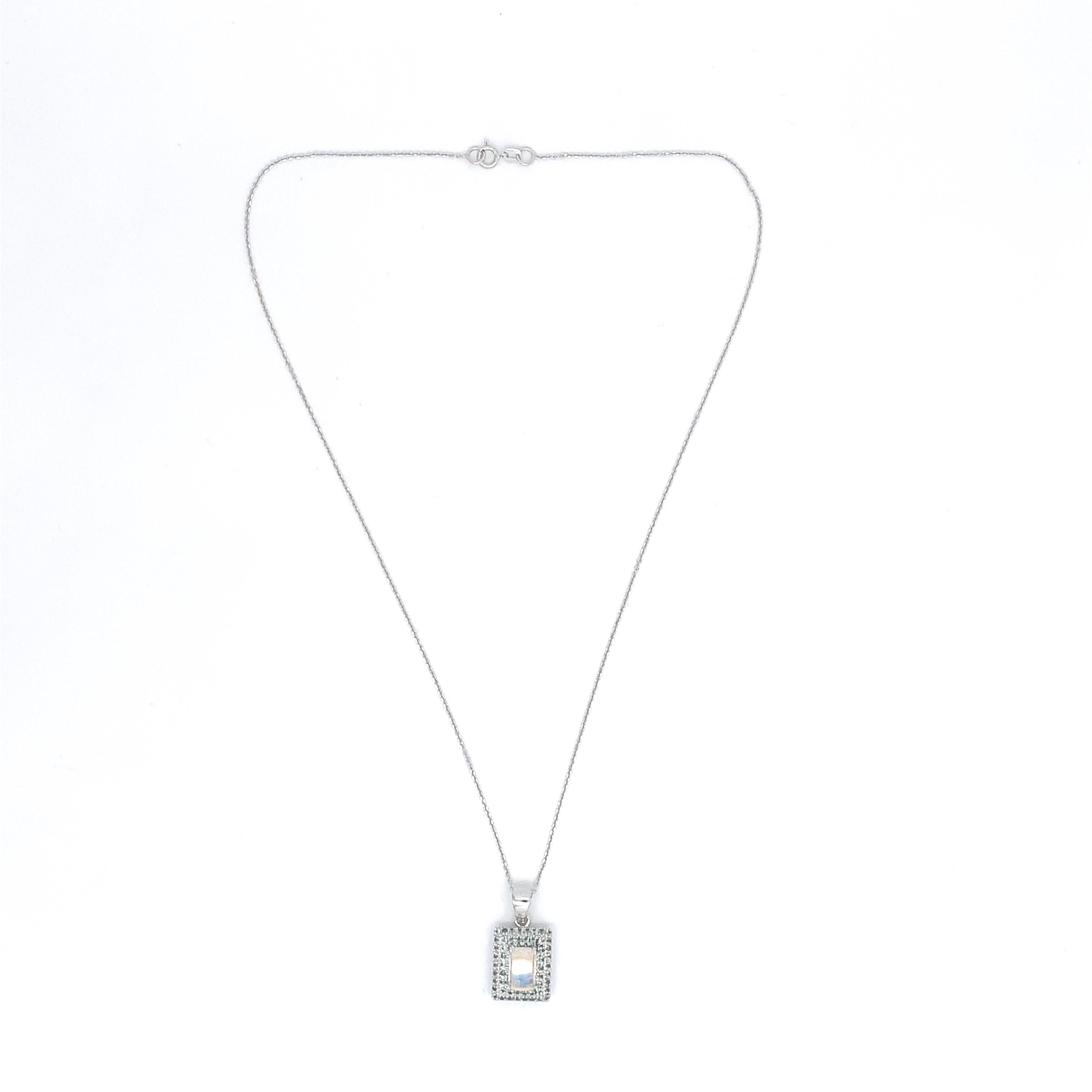 Pendentif en or blanc 14 carats pierre de lune arc-en-ciel et saphir sarcelle orné d'une pierre de lune taillée en coussin pesant 2,5 carats. La pierre de lune est transparente/claire avec des reflets bleus et mesure environ 8,8 mm x 5,5 mm. Il y a