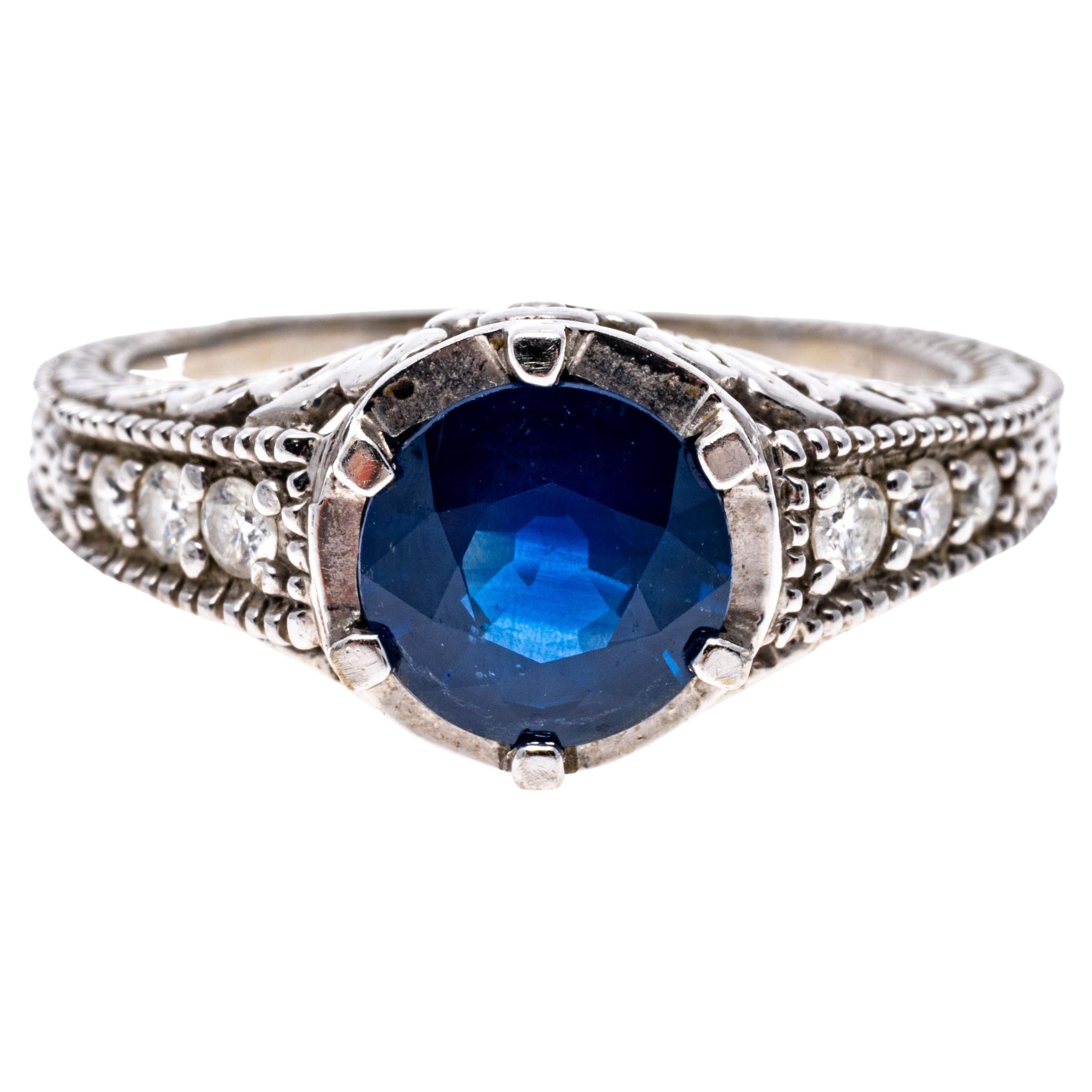 14k White Gold Round Blue Sapphire, 1.44 CTS And Diamond Ring.
This impressive ring has a round faceted sapphire center stone of navy blue color, 1.44 CTS with a white gold halo, flanked by near colorless round brilliant cut diamonds, approximately