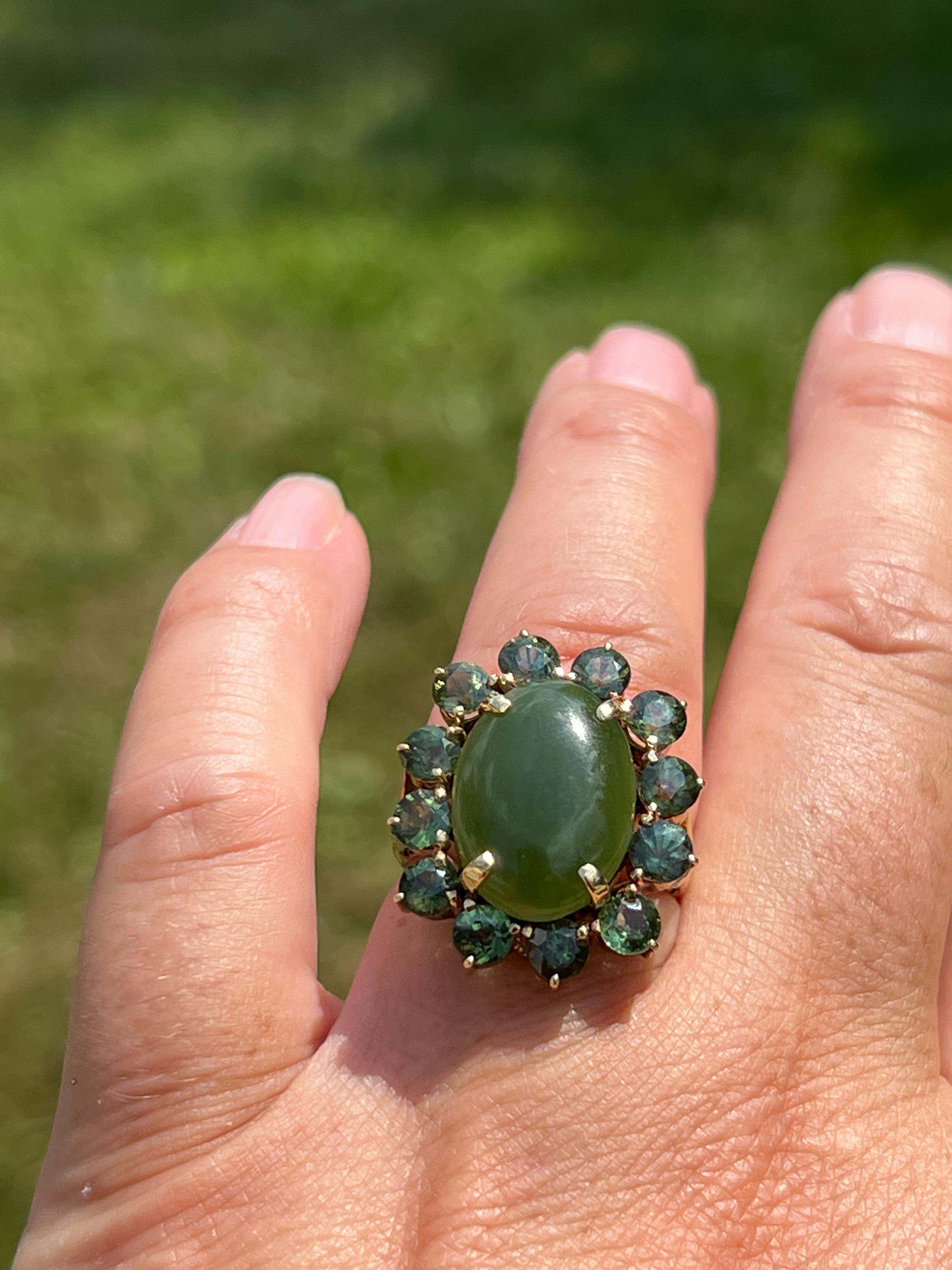 One 1960s 14 karat yellow gold cocktail ring set with one oval nephrite jade approximately 6.33 carats and twelve round green sapphires approximately 3.36 total carat weight. This ring weighs 7.76 grams, is a size 5.75 and is sizeable however this