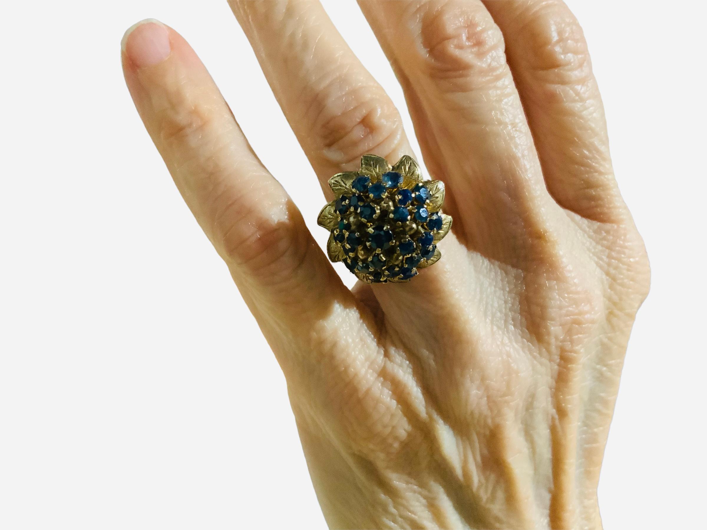 This is a 14K yellow gold blue sapphires dome cocktail ring. Its weight is approximately 6.4 g. There are 26 round cut sapphires in cluster in prong setting. A wreath of engraved gold leaves adorn the base of the dome of sapphires and enhance this