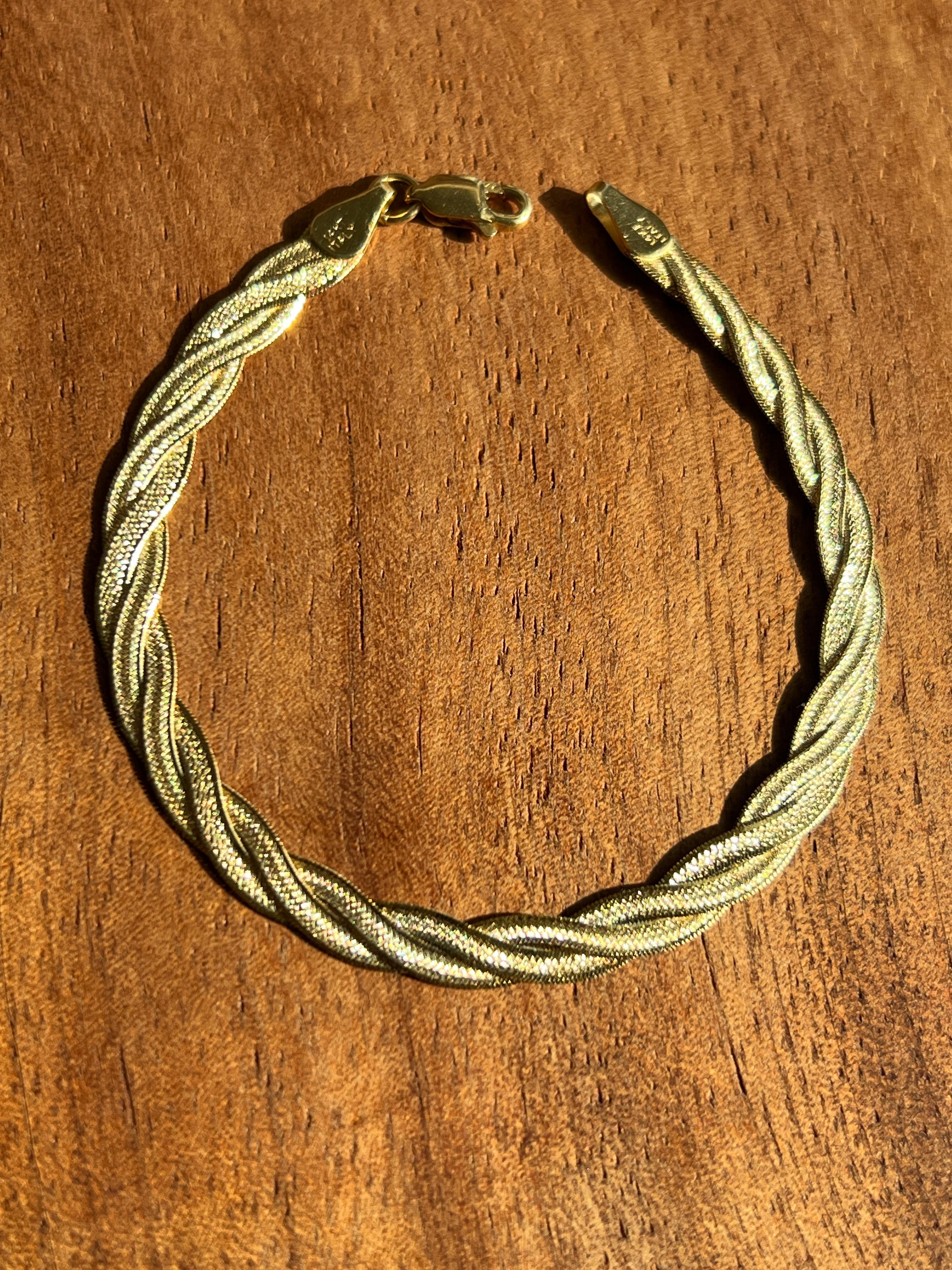 This 14-karat yellow gold braided herringbone chain bracelet combines elegance and versatility in a timeless design. Measuring 7 inches in length and 1/8 inch in width, it is lightweight at 4 grams yet substantial enough to make a statement on its