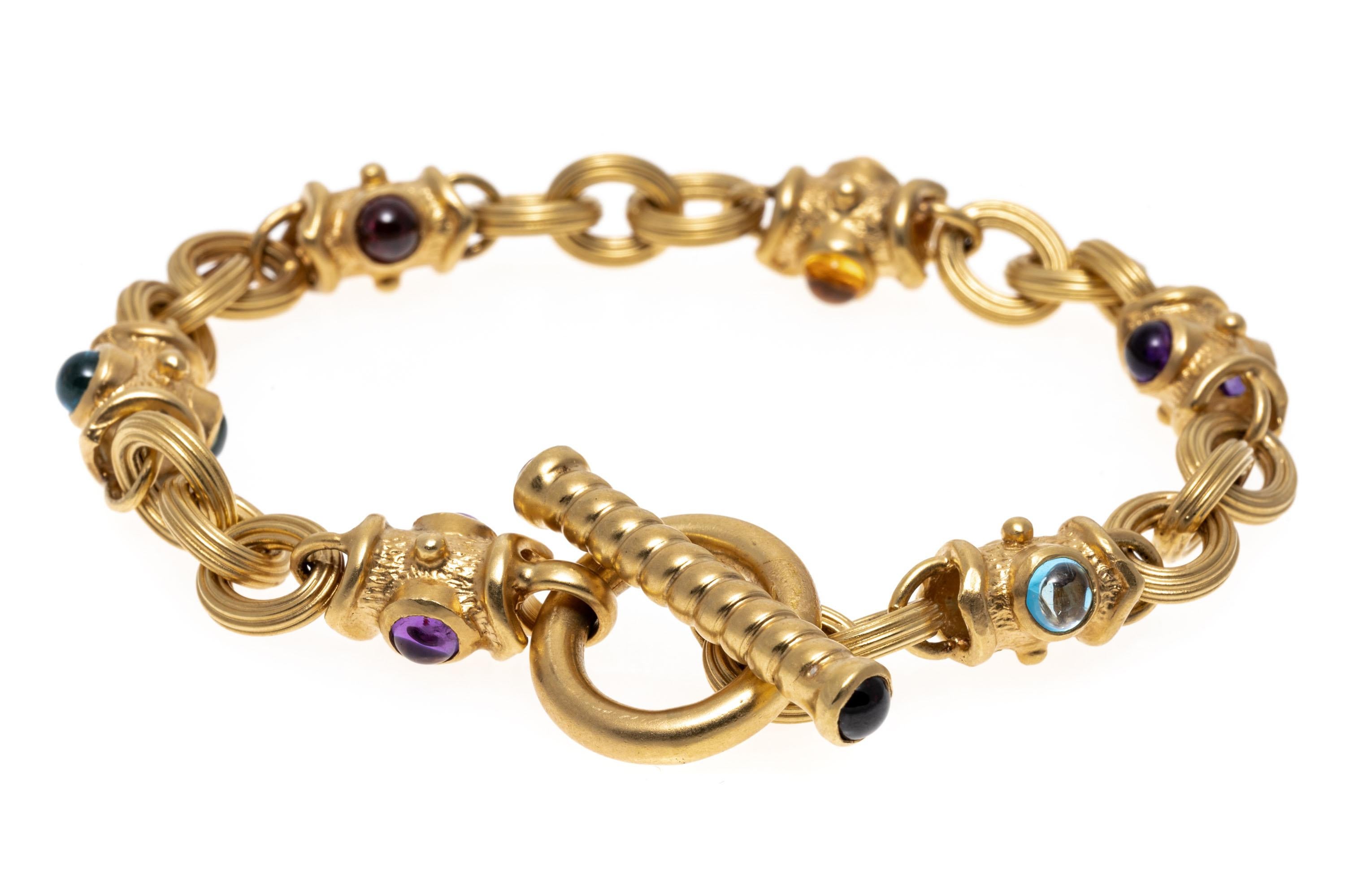 14k yellow gold bracelet. This classic bracelet is a matte finished, ribbed oval link, set with alternating Byzantine style links, set with round cabachon blue topaz, amethyst, citrine and garnet stones. The bracelet has a toggle style clasp.
Marks:
