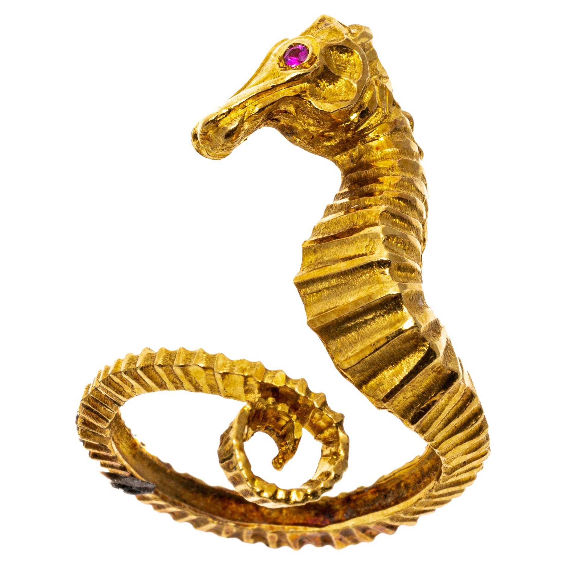 14k yellow gold ring. This whimsical, yellow gold ring has a center, figural coiled sea horse, facing to the left with a ribbed body and a round faceted synthetic red ruby set eye. The ribbing of the body is extended around the wrap-around shank,
