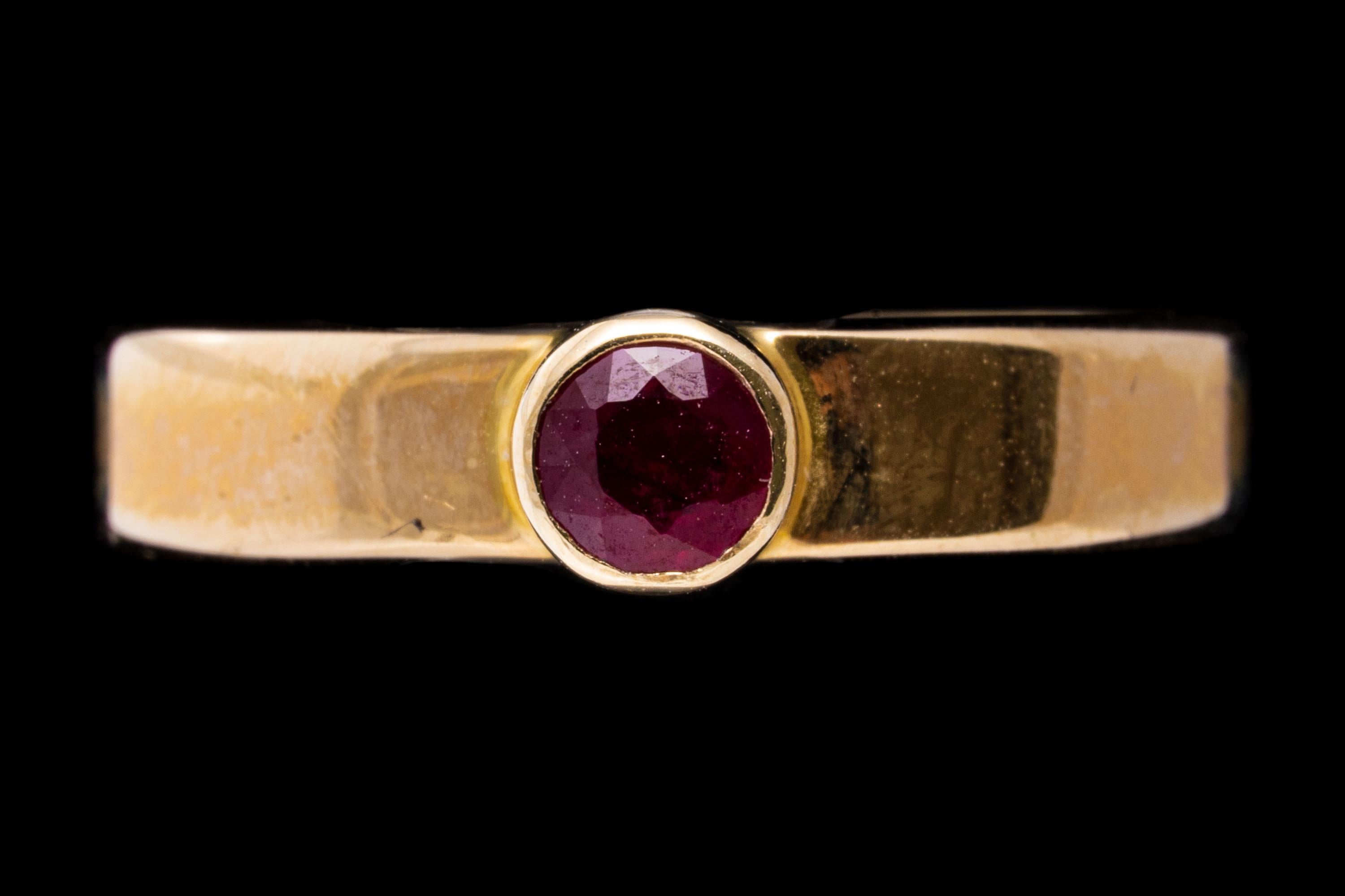 14k yellow gold ring. This simple contemporary band ring is set with a center round faceted, medium to dark pinkish red color ruby, approximately 0.18 CTS, set with a high bezel and finished with a high polished finish.
Marks: 14k 
Dimensions: 3/16