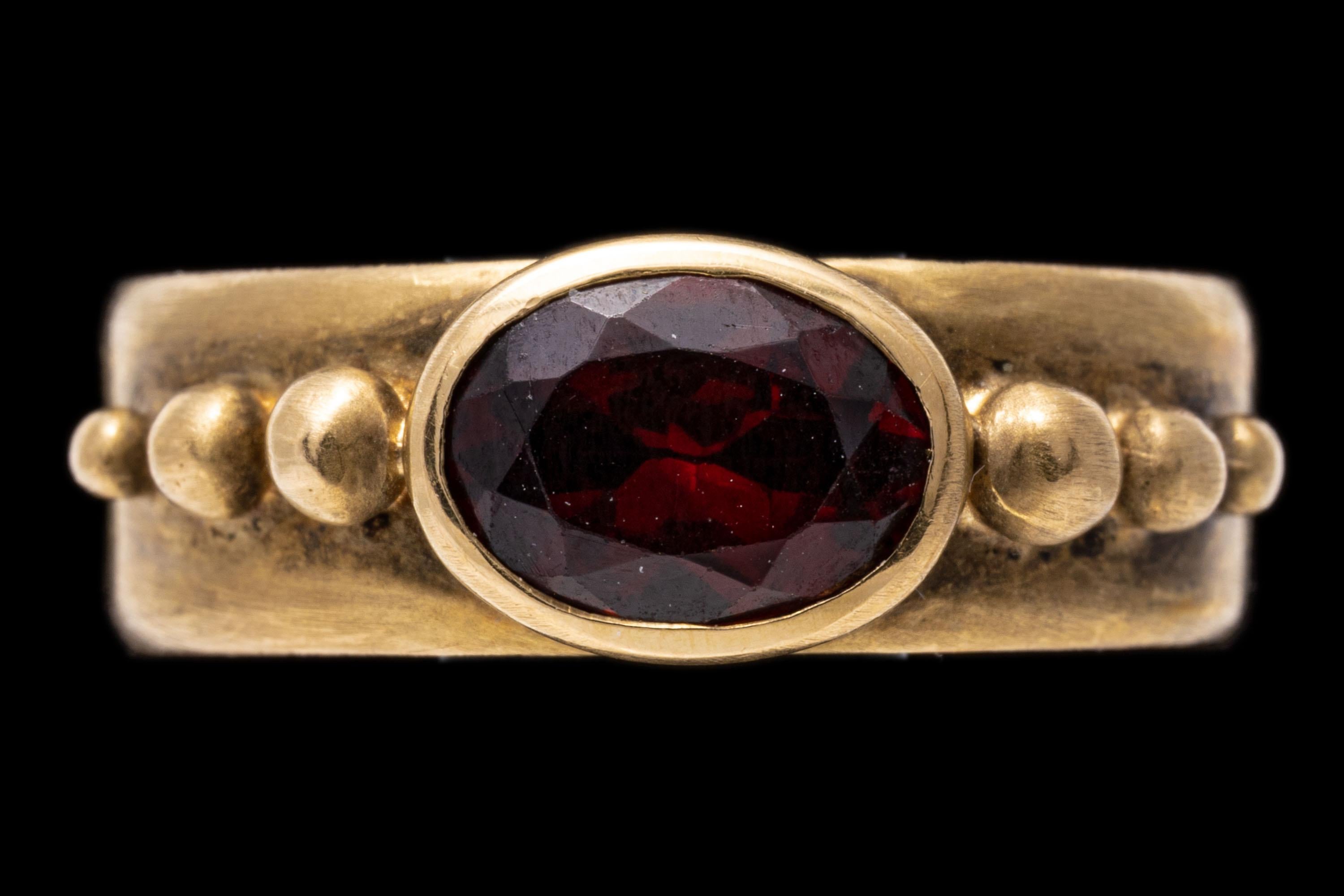14k yellow gold ring. This stylish ring has a horizontal oval faceted, medium to dark burgundy color garnet, approximately 1.30 CTS and bezel set into a wide, matte burnished band and flanked by graduated ball sides.
Marks: 14k 
Dimensions: 1/4