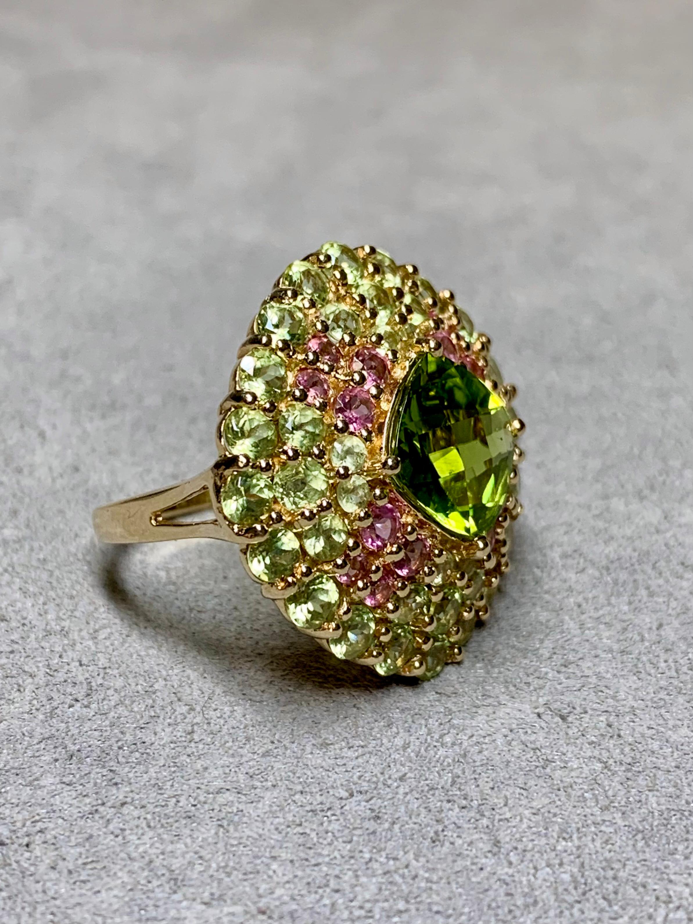
A bold, contemporary cocktail ring done in 14K yellow gold and centered by a deep greed natural cushion cut peridot weighing approximately 3.50ctw. Surrounding the center stone are numerous lighter colored peridot as well as soft, natural pink