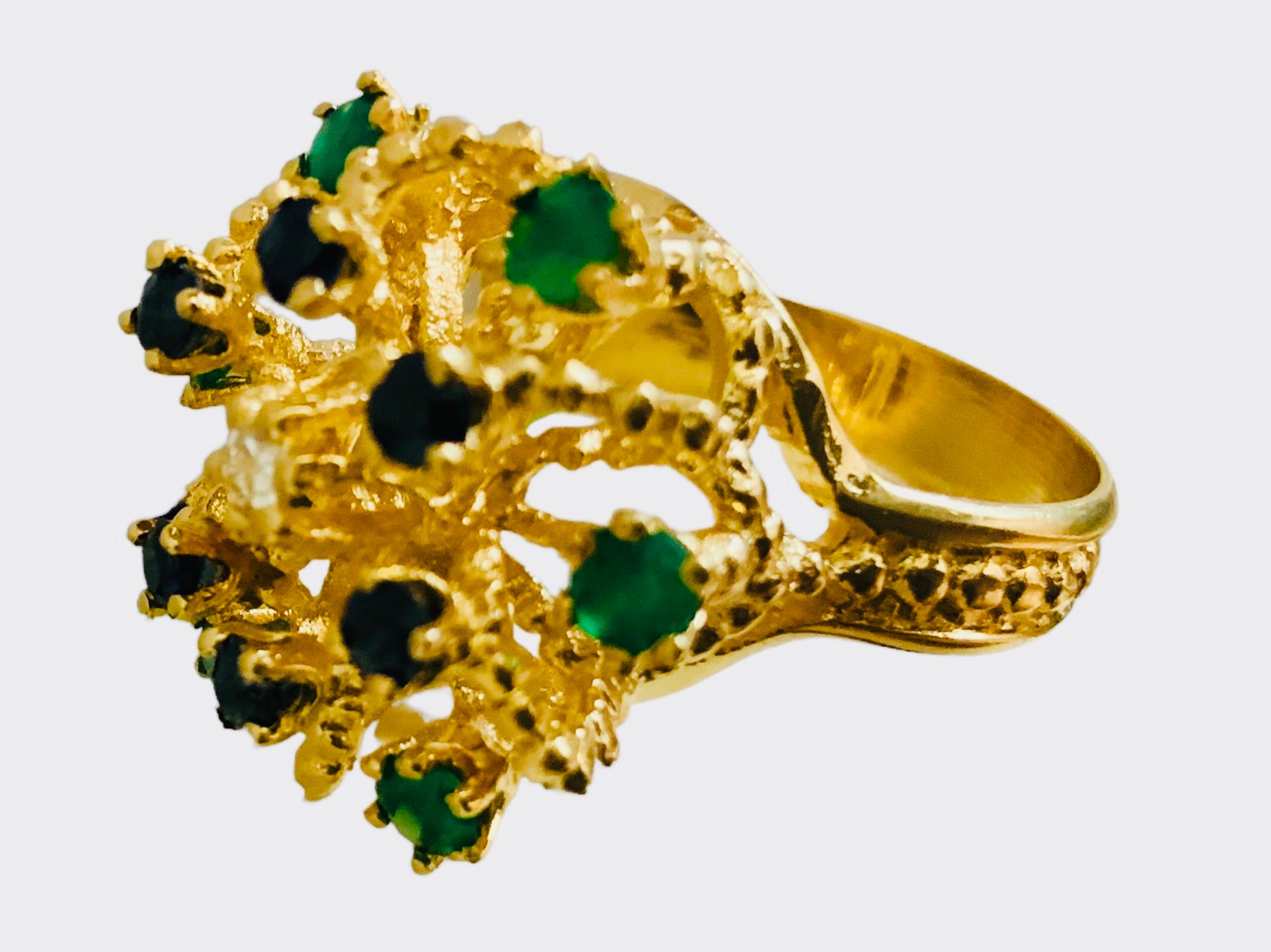 This is a 14K yellow gold Diamond, Emeralds and Sapphires cocktail ring. It depicts a crown like setting with two tiers. The lower tier is enhanced by six round tiny emeralds in prong setting and the higher tier is adorned by six round tiny
