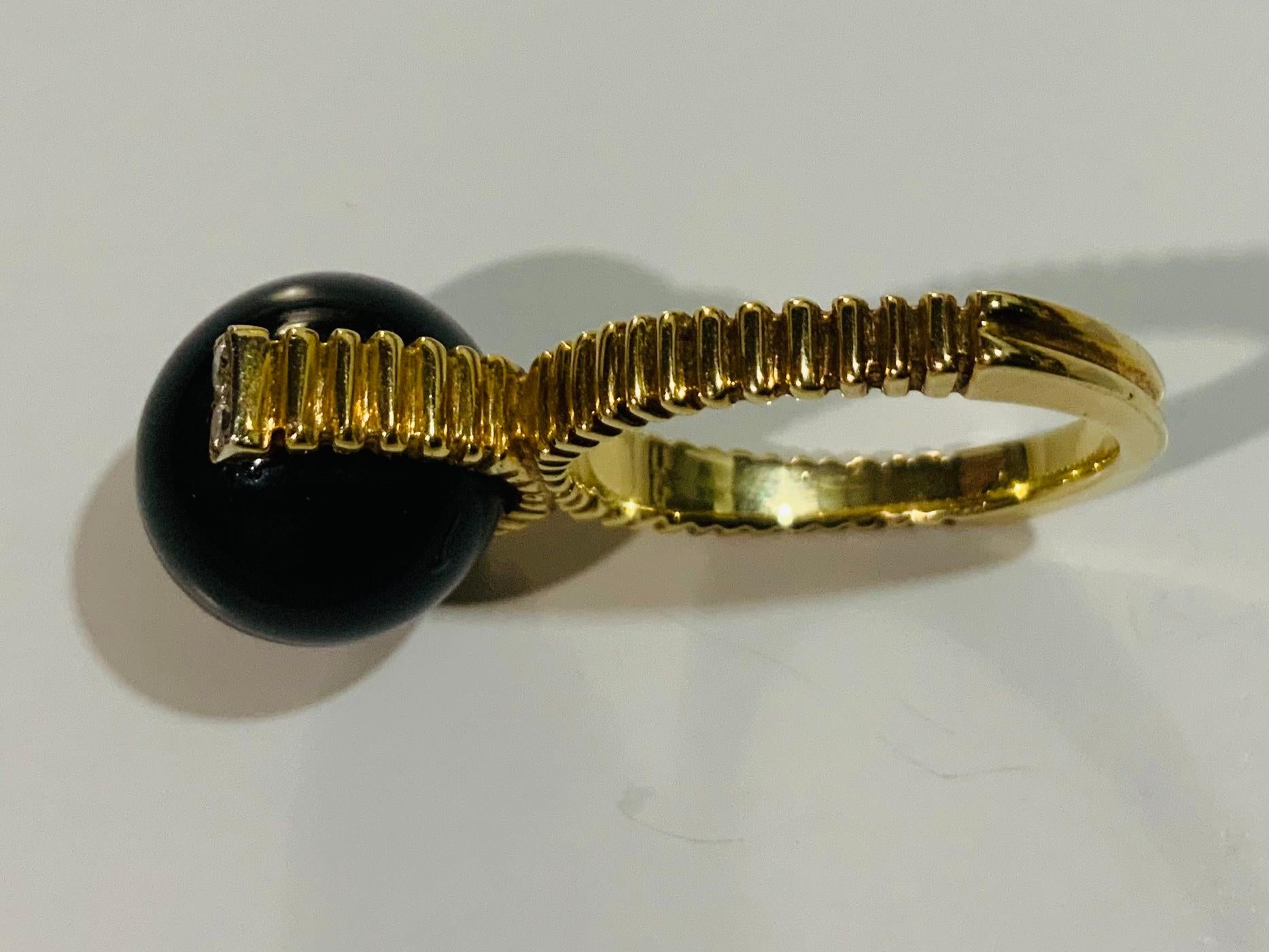This is a 14K white gold, diamonds and black stone cocktail ring. It depicts a black stone sphere supported by a yellow gold ribbed concave trough arc enhanced by four round tiny diamonds in pave setting at each top ends. Also, both shanks of the
