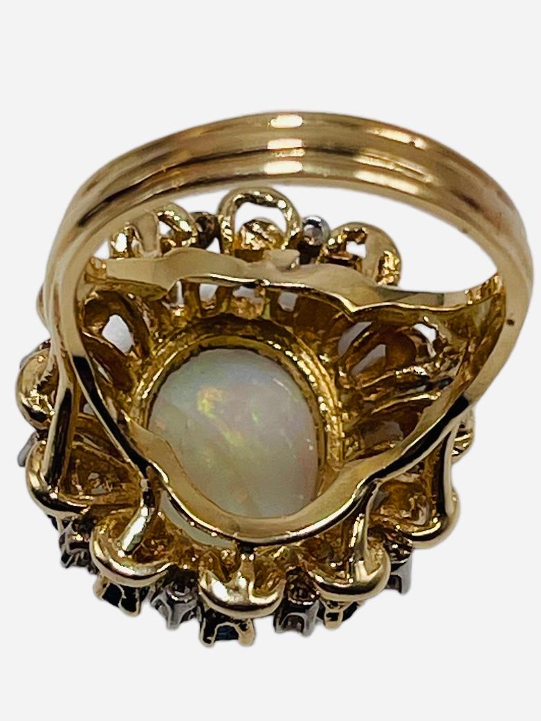 This is a 14K yellow gold diamonds, opal and sapphires cocktail ring. The ring forms a flower. This one is made of an external large halo of twelve tiny round cut diamonds in prong setting followed by a small halo of twelve round cut sapphires in