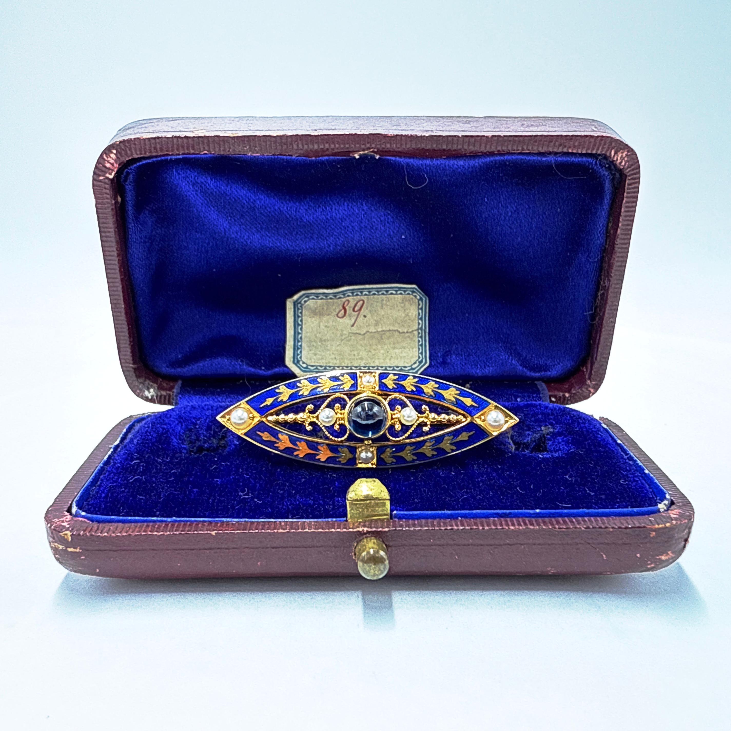 This piece is an Edwardian early 20th century brooch and pendant combination crafted in 14K yellow gold. The design features a marquise-shaped openwork form, accented with blue enamel, seed pearls, and a cabochon-cut sapphire in a central bezel