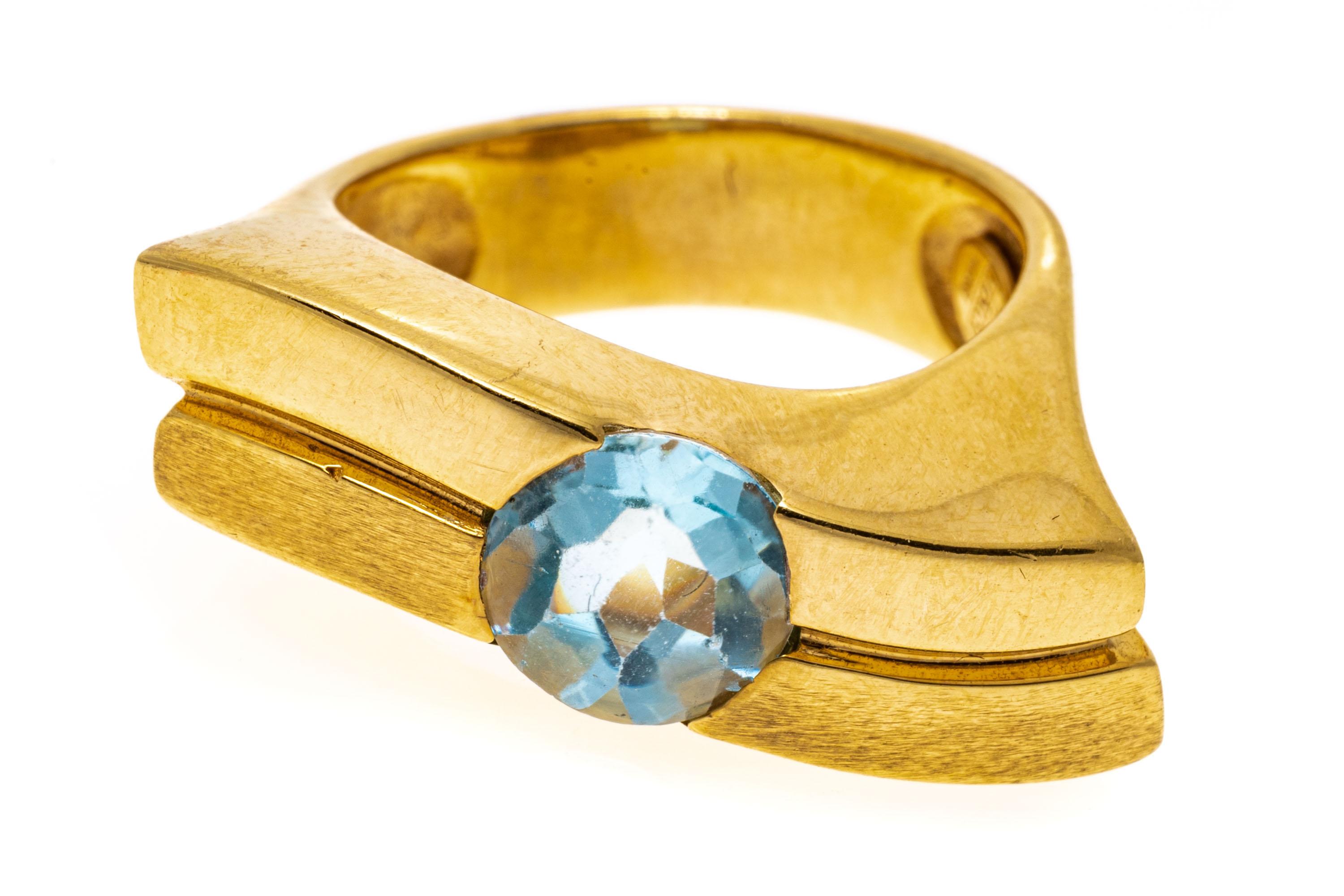 14k yellow gold ring. This unusual ring has a round, half faceted and half cabachon cut, light blue color blue topaz, center flush set into a wide, elongated contemporary matte and high polished lined waved top, decorated with wide, split