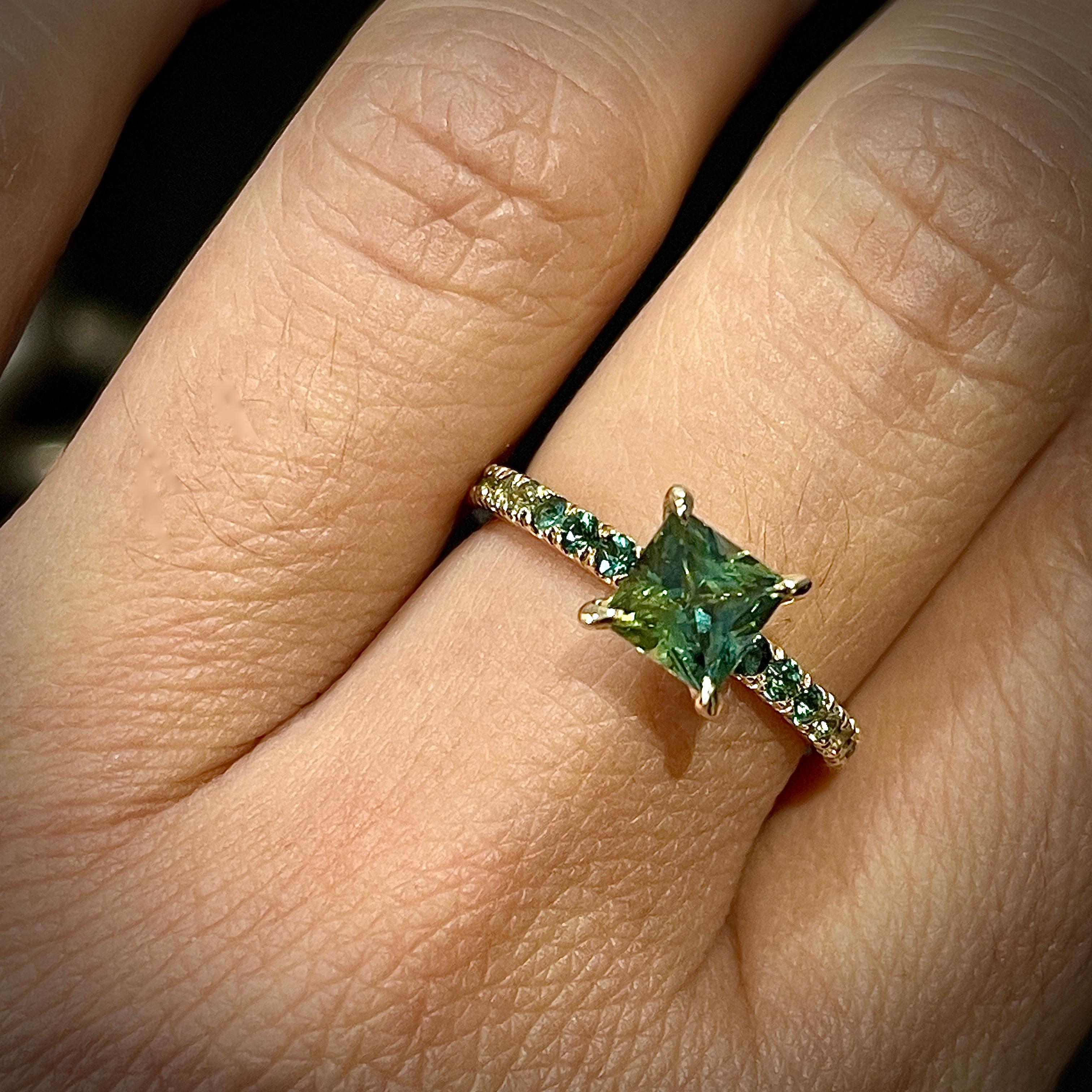 Rehaussez vos fiançailles avec une bague vraiment captivante et unique. Cette pièce exquise présente un magnifique saphir vert de 1,31 carat de taille princesse, délicatement serti dans un anneau d'or jaune 14ct. Les côtés de l'anneau sont ornés de