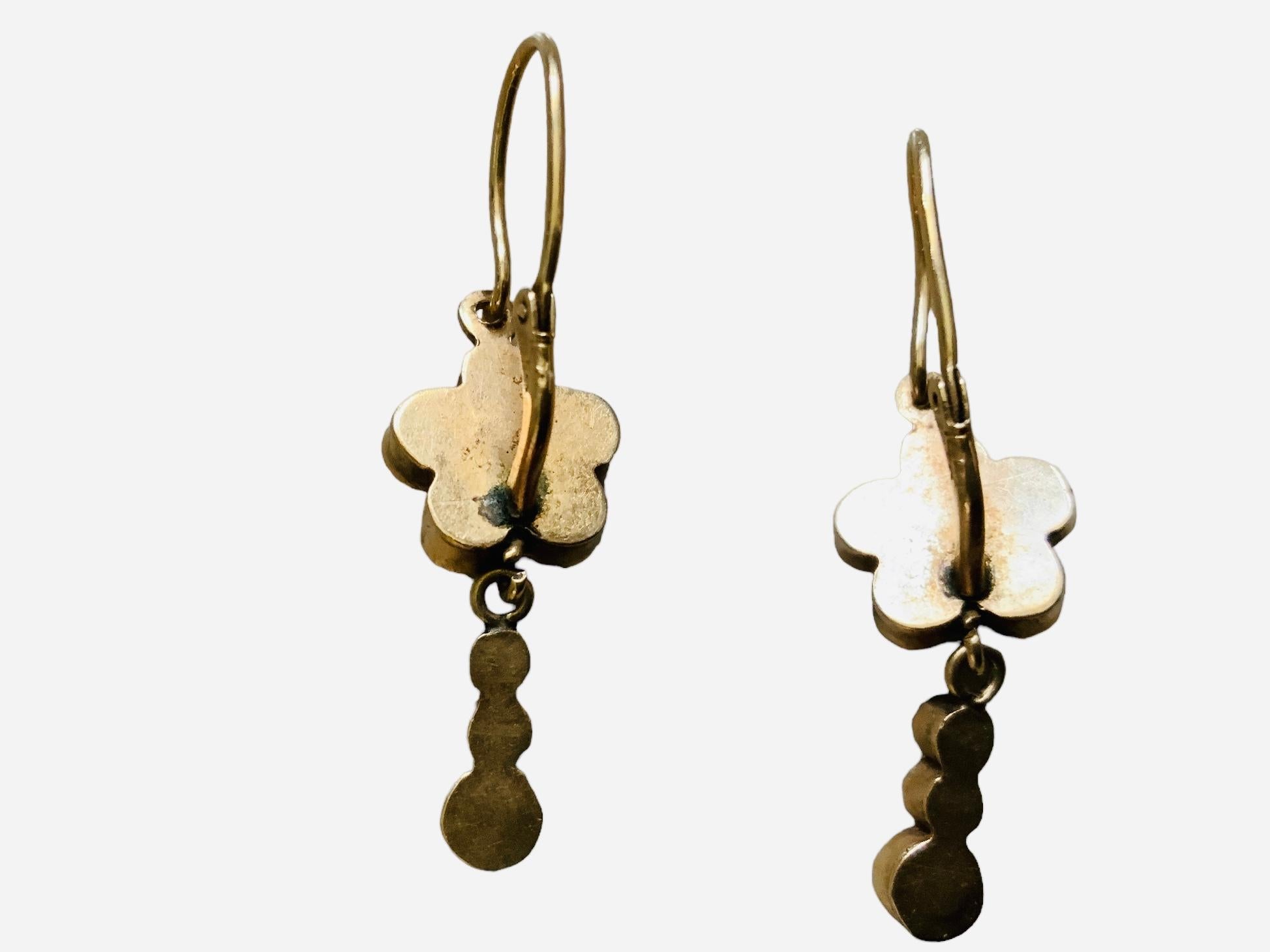 This is a Pair of 14K yellow gold Drops Earrings. It depicts a flower made of six tiny cylindrical cases that contain light green faceted glasses mounted in 14K gold bezel setting. From this flower hang three tiny cylinders with the same glass. Both