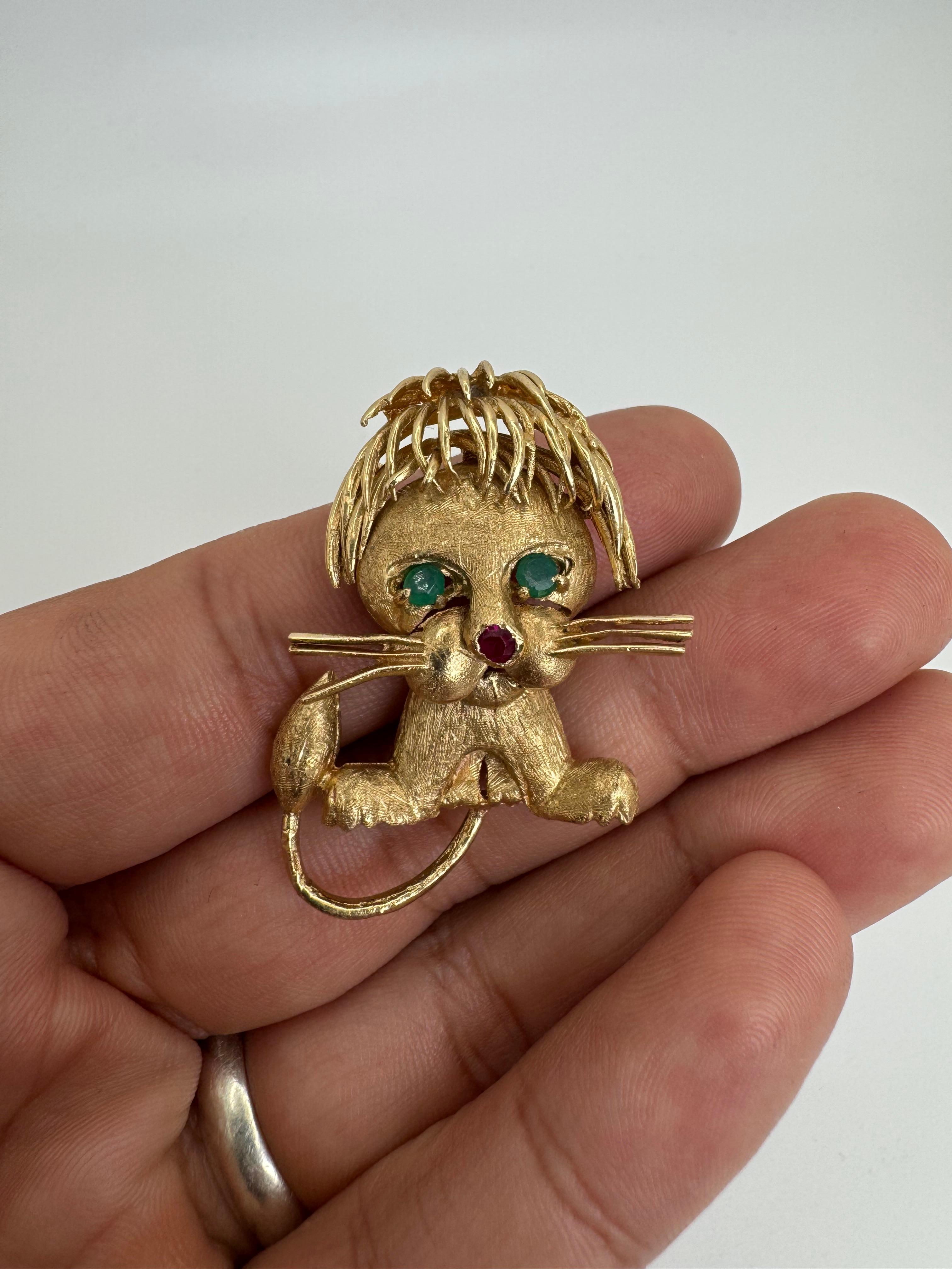 Este encantador broche de león vintage está fabricado en oro amarillo de 14 quilates con una rica textura, y muestra un diseño juguetón y lleno de carácter. La expresiva cara del león presenta vibrantes cabujones de esmeralda como ojos y un rubí