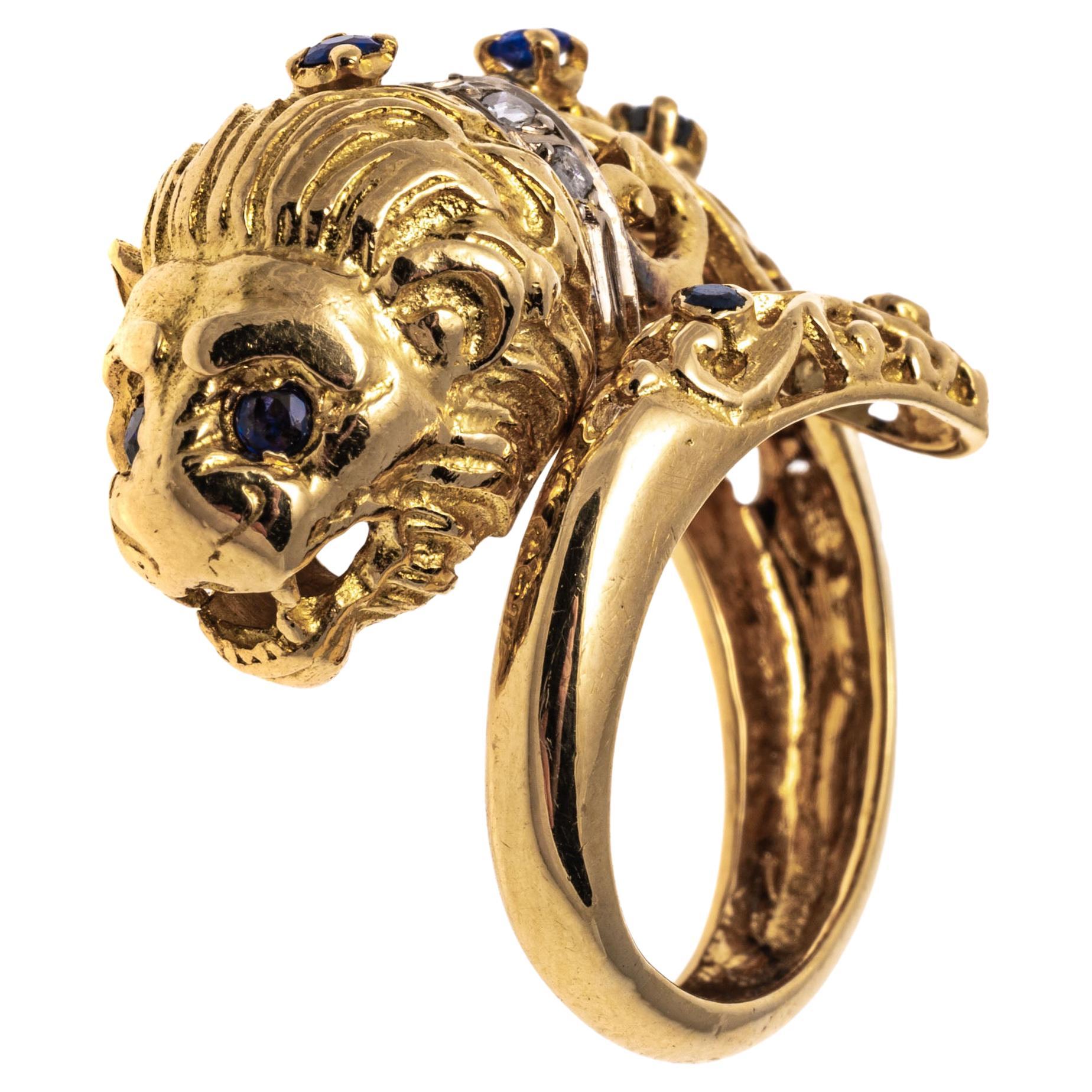 14k yellow gold ring. This striking, ornate yellow gold bypass ring has a center high polished finished, figural lions head, with round faceted sapphires set in the eyes and studded down the backs, approximately 0.25 TCW and decorated with a collar