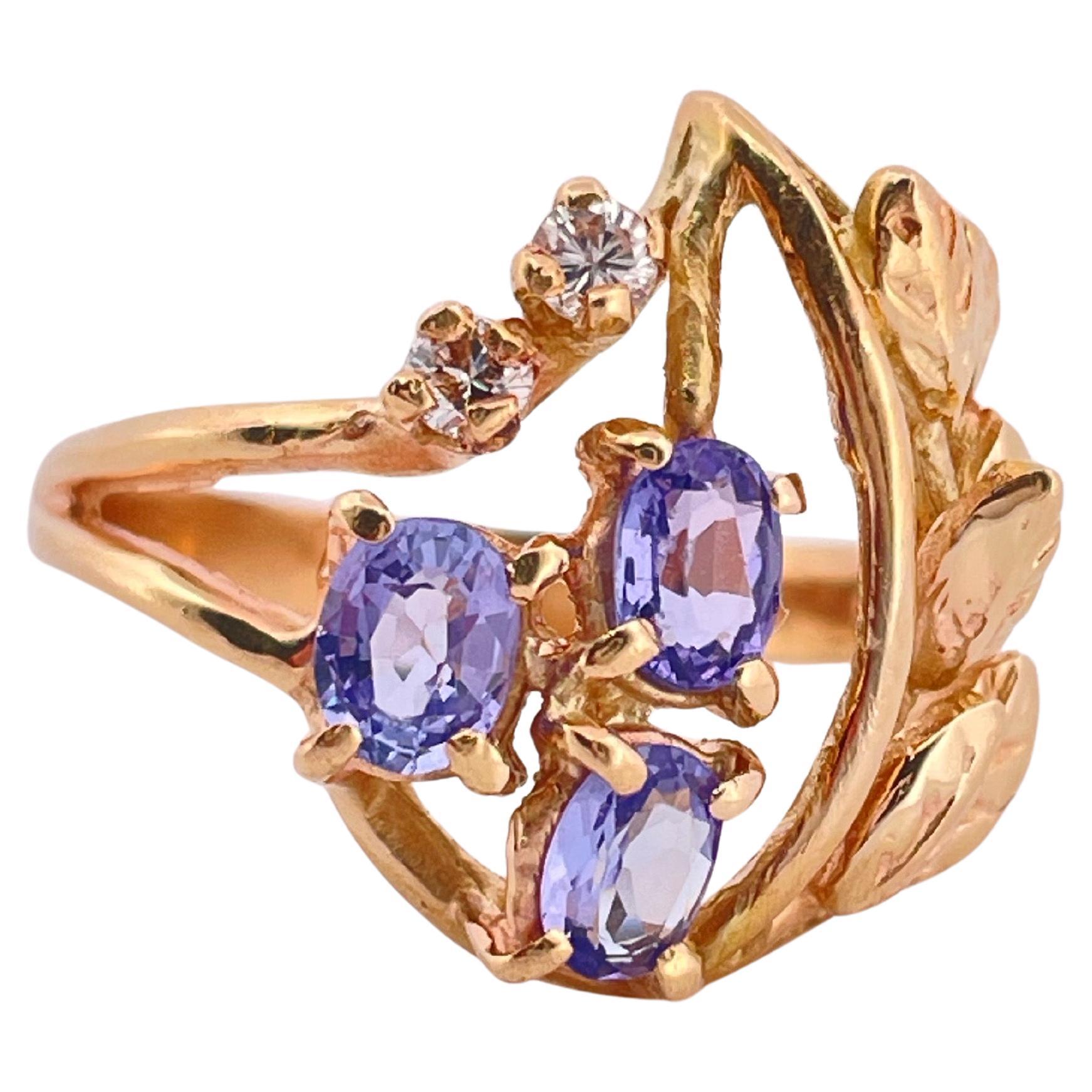 Dieser elegante Ring aus 14-karätigem Gelbgold besticht durch sein floral inspiriertes Design, das mit leuchtenden Tansaniten und funkelnden Diamanten verziert ist. Drei Tansanite im Ovalschliff, die für ihren verführerischen violettblauen Farbton