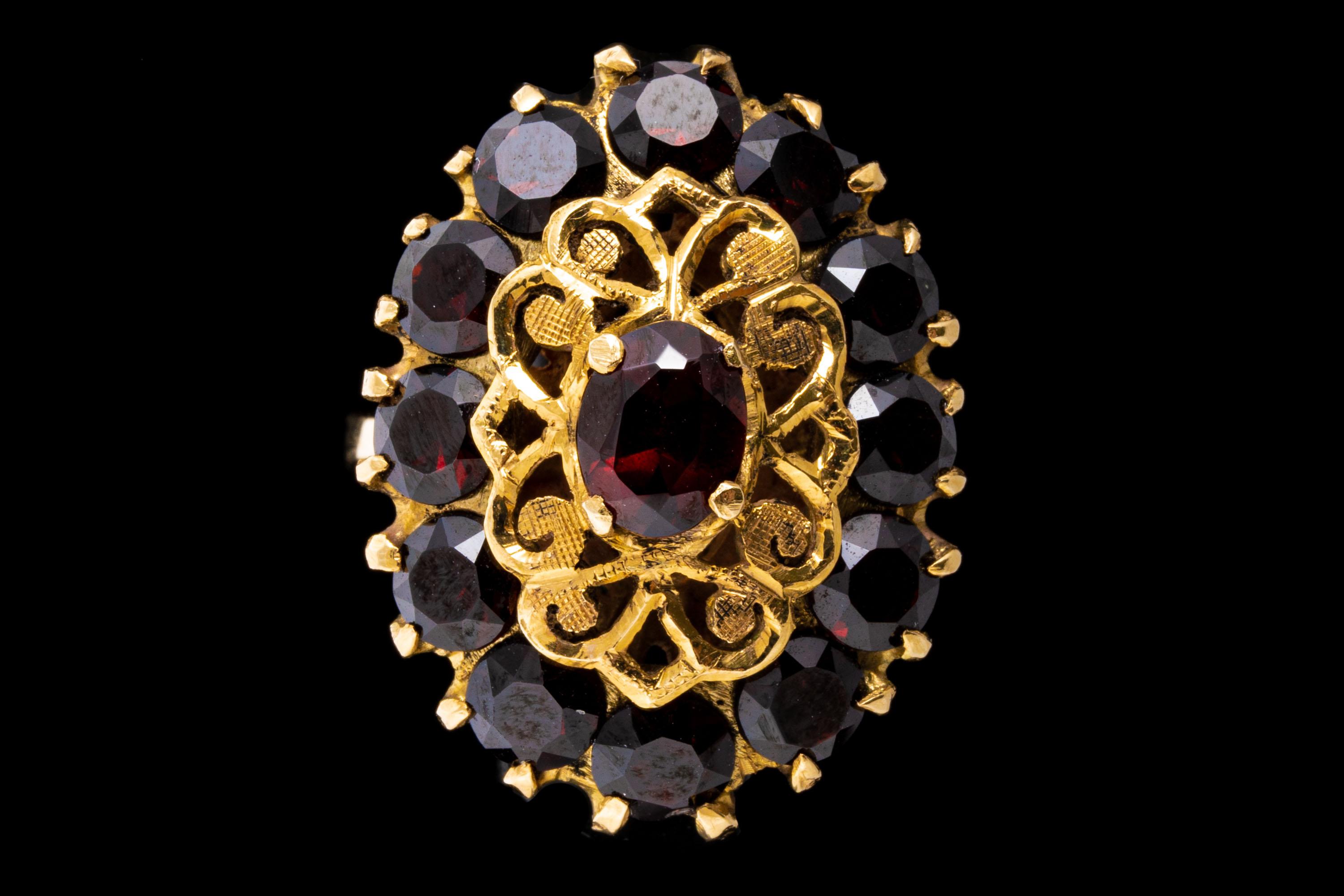 18k yellow gold ring. This vintage retro ring is an elongated oval motif cocktail style, decorated with an oval faceted, dark burgundy color center, approximately 0.49 CTS, surrounded by a pierced filigree borderand framed by a halo of round