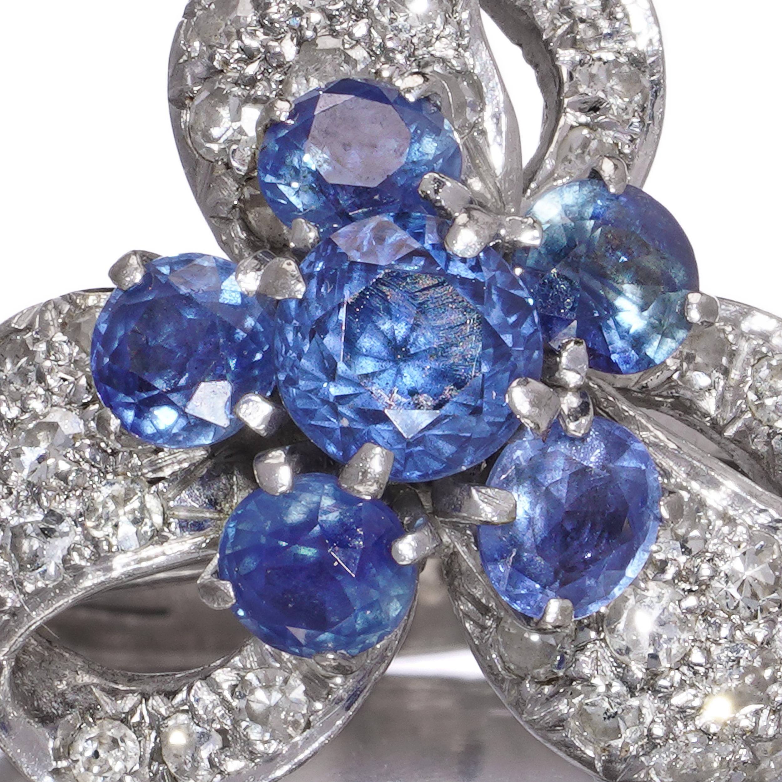 A striking and substantial ring crafted in 14kt white gold, featuring a sculptural, ribbon-like cluster design set with brilliant diamonds and rich blue sapphires. The dynamic composition resembles a folded bow or floral motif, with a strong