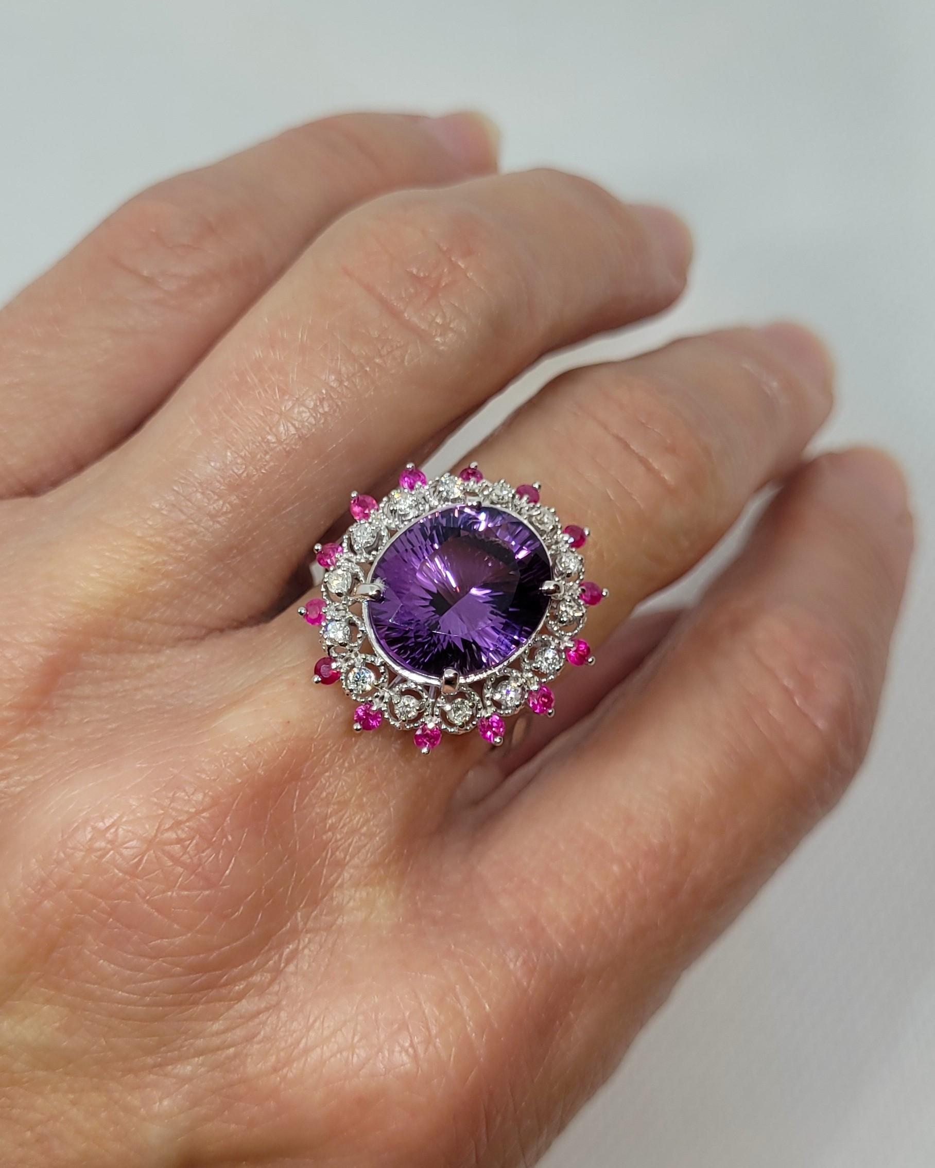 Cette magnifique bague en or blanc 14kt est ornée d'une améthyste ovale de 6,50ct, connue pour son excellente taille et sa couleur violette vive, mesurant 13,25mm x 11,11mm. La belle pierre centrale est entourée d'un halo de diamants et de rubis. La