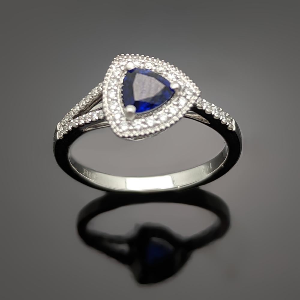 A unique 14kt white gold ring featuring a trillion cut blue sapphire with an estimated weight of 0.55ct featuring a halo of diamonds, split-shank on one side, and diamonds down the shoulders. Diamonds are estimated 0.20cttw.  Estimated weight of