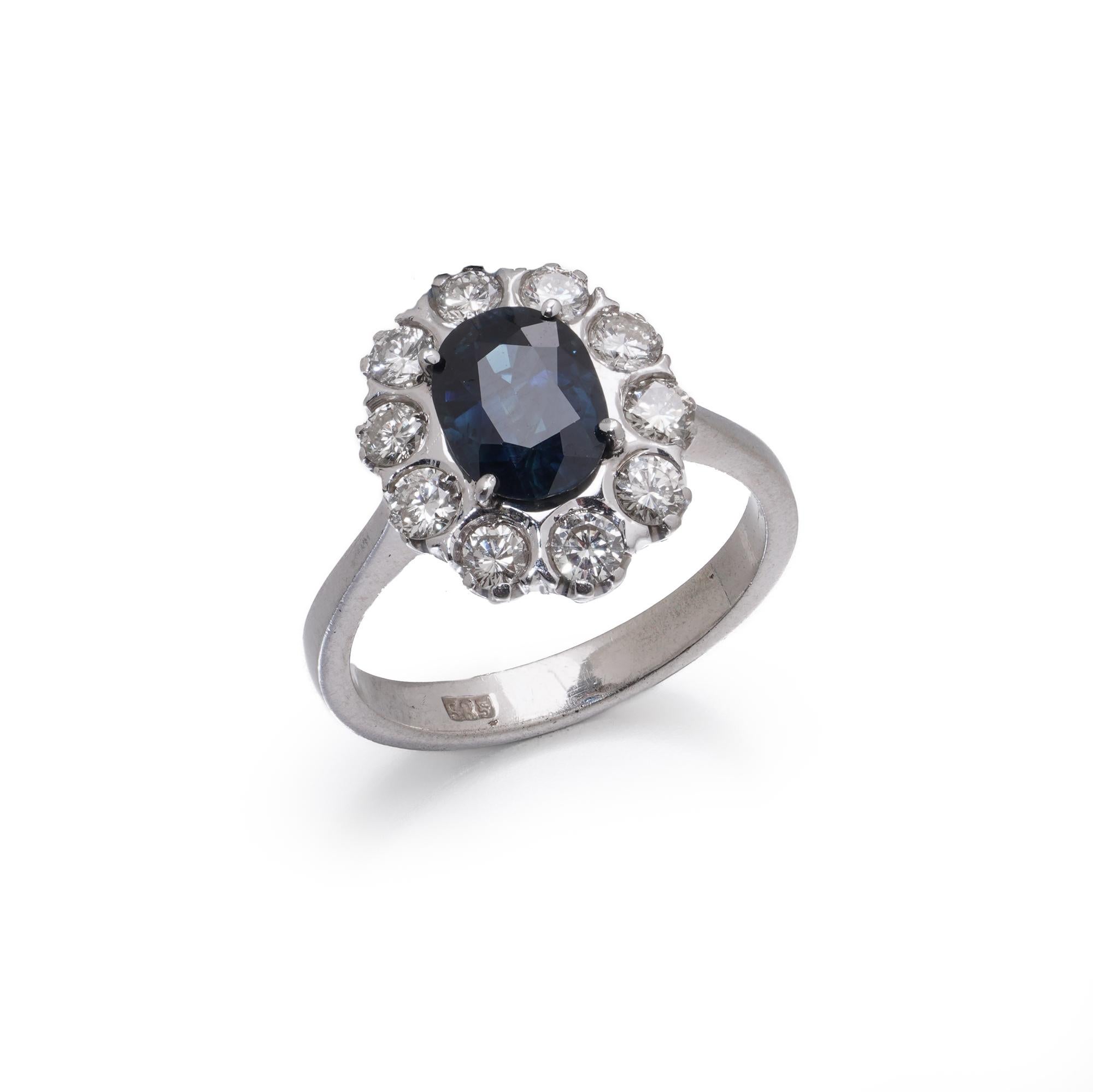 Ein auffälliger Vintage-Ring aus 14-karätigem Weißgold mit einem ovalen, grünlich-blauen Saphir im Mischschliff, umgeben von zehn runden natürlichen Diamanten im Brillantschliff in einem klassischen Halo-Design. Der Ring ist durch das World