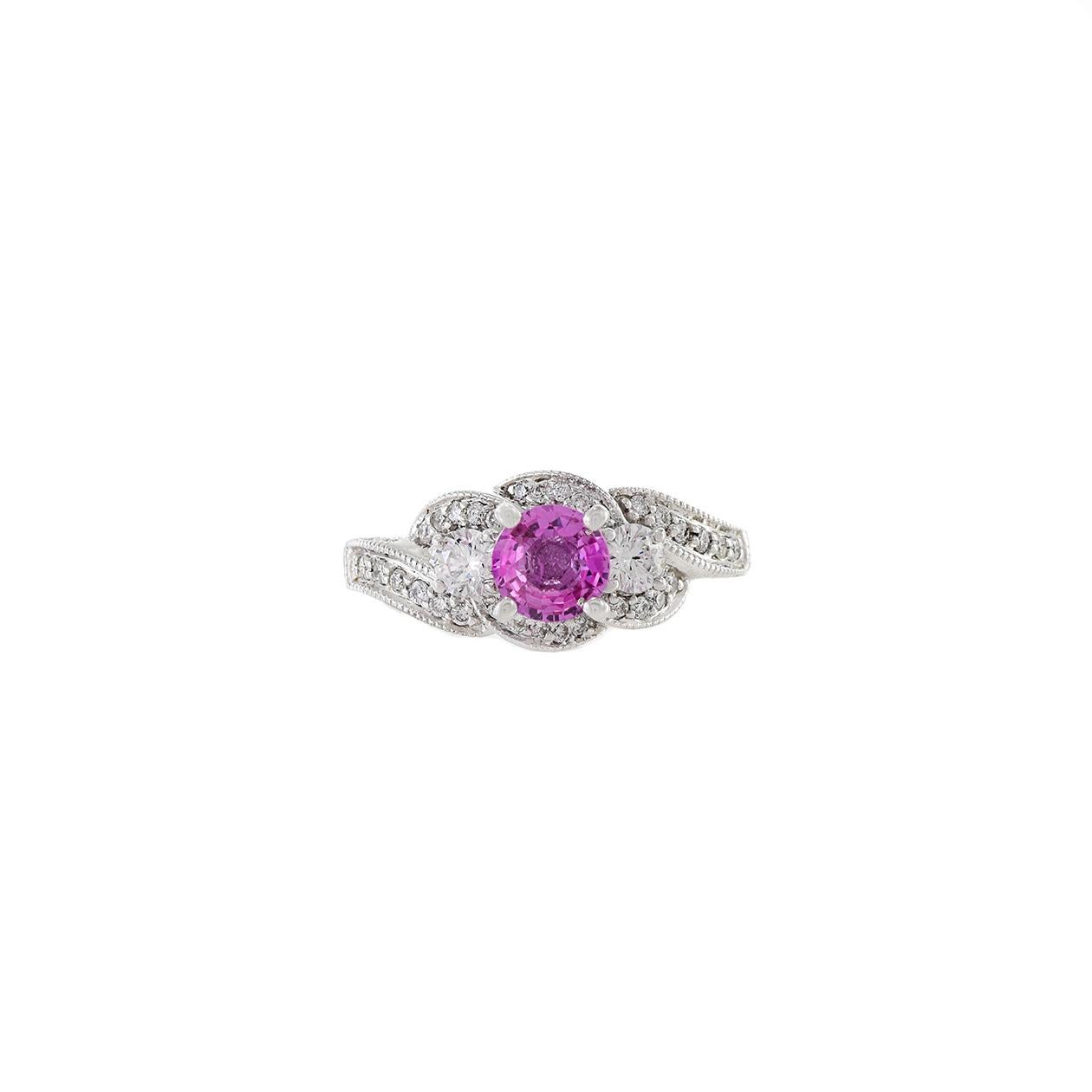 This 14KT white gold ring is a stunning and elegant piece of jewelry, featuring a genuine round-cut Ceylon pink sapphire with a weight of 1.02 carats at its center. The pink sapphire is beautifully surrounded by a swirling design of round-cut