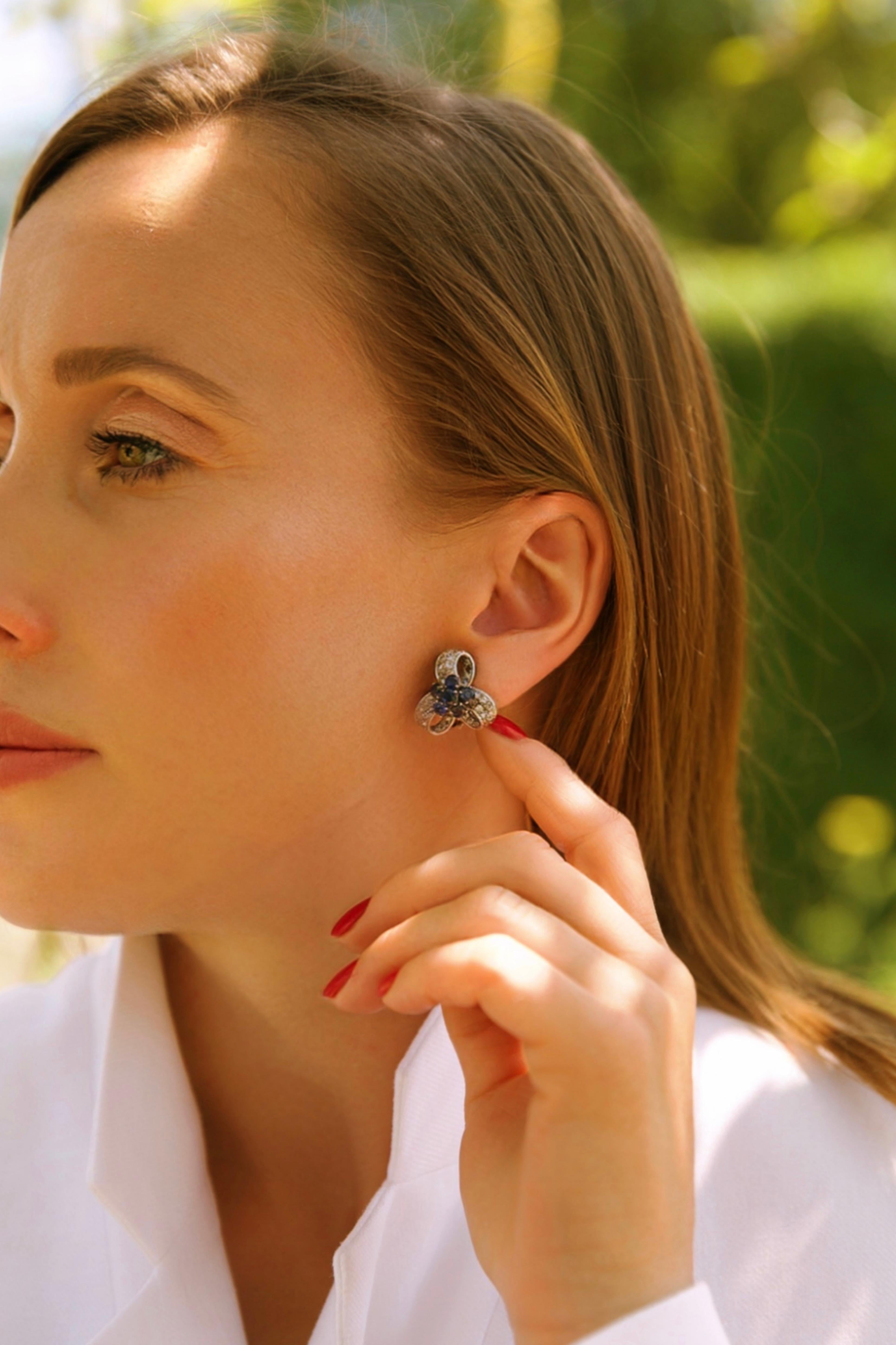 An elegant pair of vintage earrings in 14kt white gold, designed in a stylised ribbon and floral motif. Each earring features a central flower-like cluster of 6 round-cut sapphires, framed by sculptural ribbon loops pavé-set with a total of 48