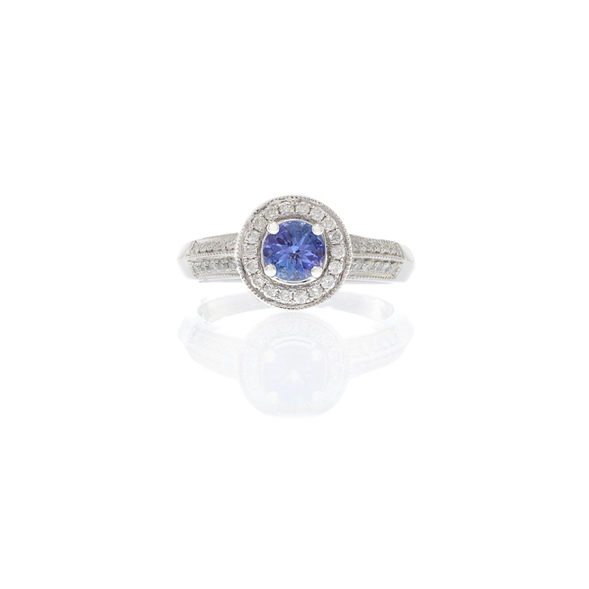 A stunning 14KT white gold ring featuring a vibrant 0.95 carat round cut tanzanite center stone, framed by a delicate halo of sparkling round cut diamonds totaling 0.15 carats. The intricate double halo design is complemented by diamond-accented