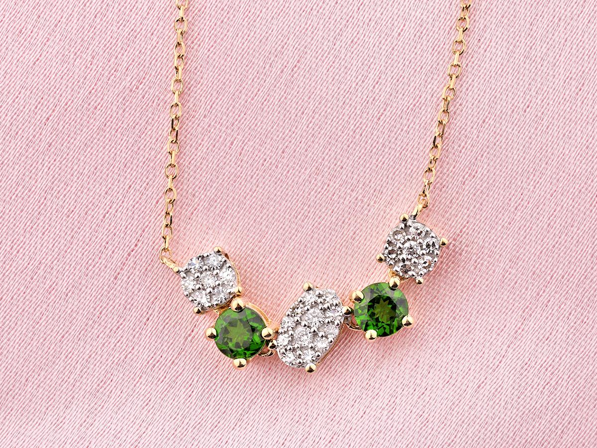 Le collier présente un design élégant et contemporain où les pierres précieuses sont disposées en forme d'arc doux ou de barre incurvée. Le motif alterne entre des pierres individuelles vertes de taille ronde et des grappes de petits diamants de