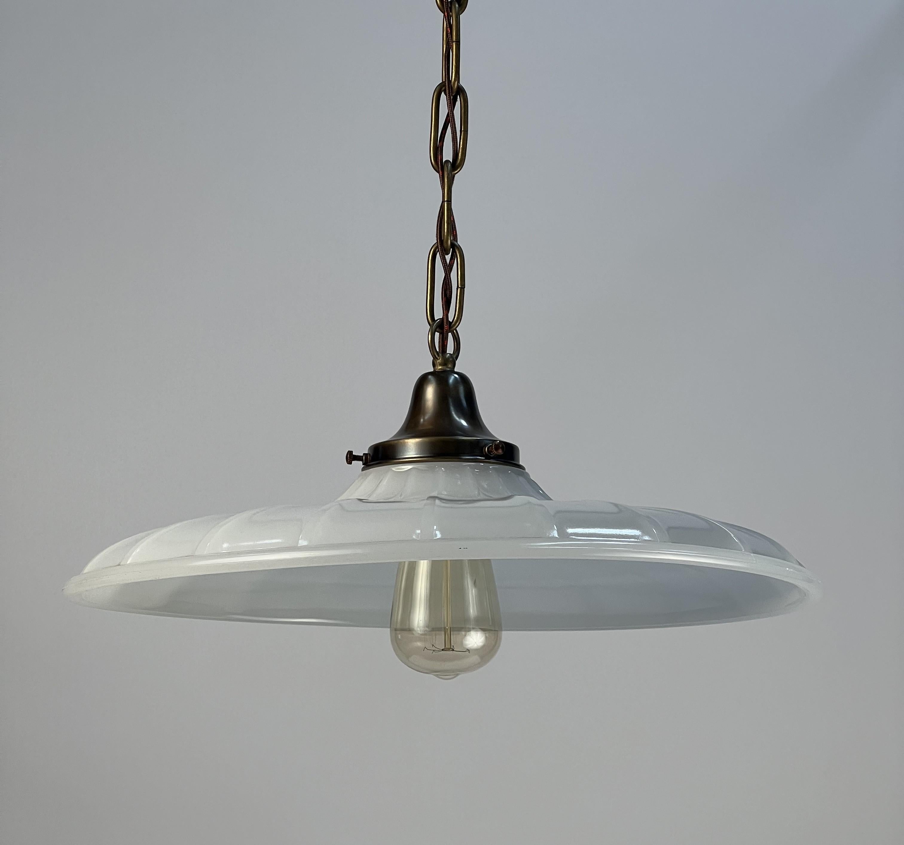 An industrial milk glass shade featuring raised ribs leading from the brass fitter to a 3/4 rounded lip. This type of glass shade would have been found in a number of industrial settings and retail stores in the early 20th century. The milk glass