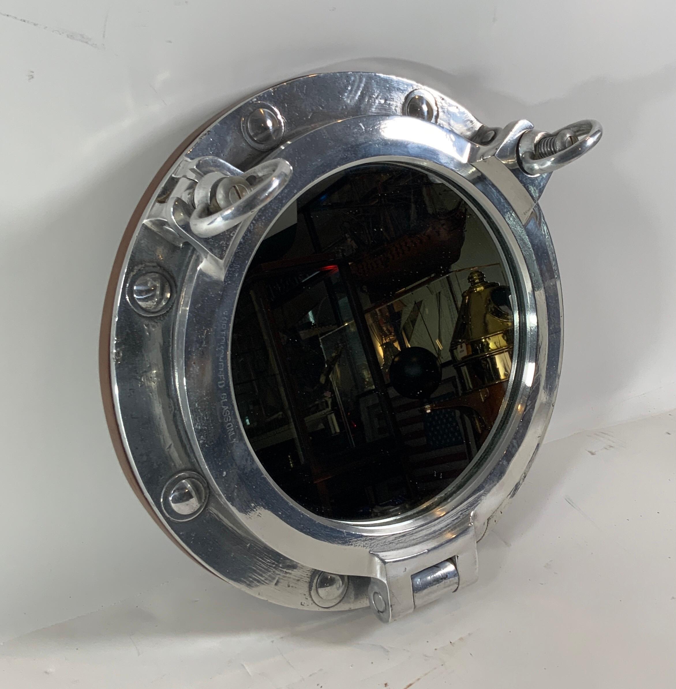 Authentic polished aluminum ship's porthole fitted with a glass mirror. Door is hinged and fitted with two dogbolts. The highly polished porthole is also fitted with a teakwood trim ring on the rear.

Overall dimensions: glass diameter: 10