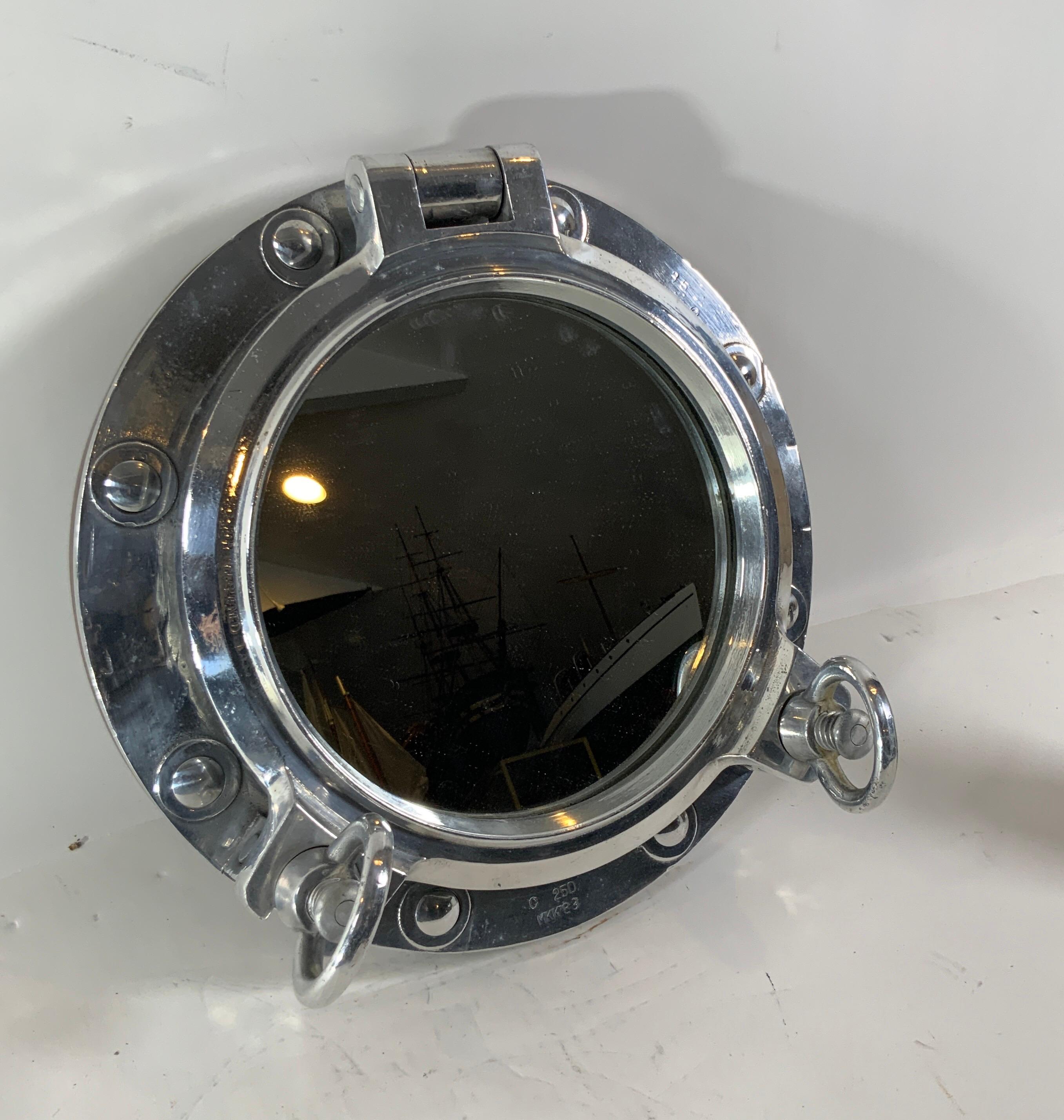 Authentic polished aluminum ship's porthole fitted with a glass mirror. Door is hinged and fitted with two dogbolts. The highly polished porthole is also fitted with a teakwood trim ring on the rear. Bolt holes are covered with caps. Substantial