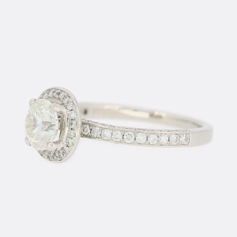 This is a platinum diamond engagement ring. The central diamond is a 0.95 carat round brilliant cut and set in a raised 4 claw mount. Surrounding the central diamond there is a halo of smaller round brilliant cut diamonds with diamonds set into the