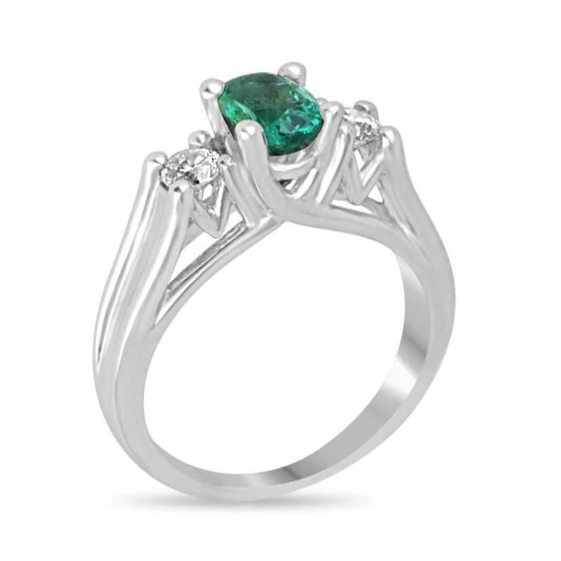 This elegant three-stone ring features a captivating oval-cut natural emerald at the center, displaying a medium rich green color and semi-transparent clarity that mesmerizes at first glance. Flanked by brilliant round-cut diamonds, the design