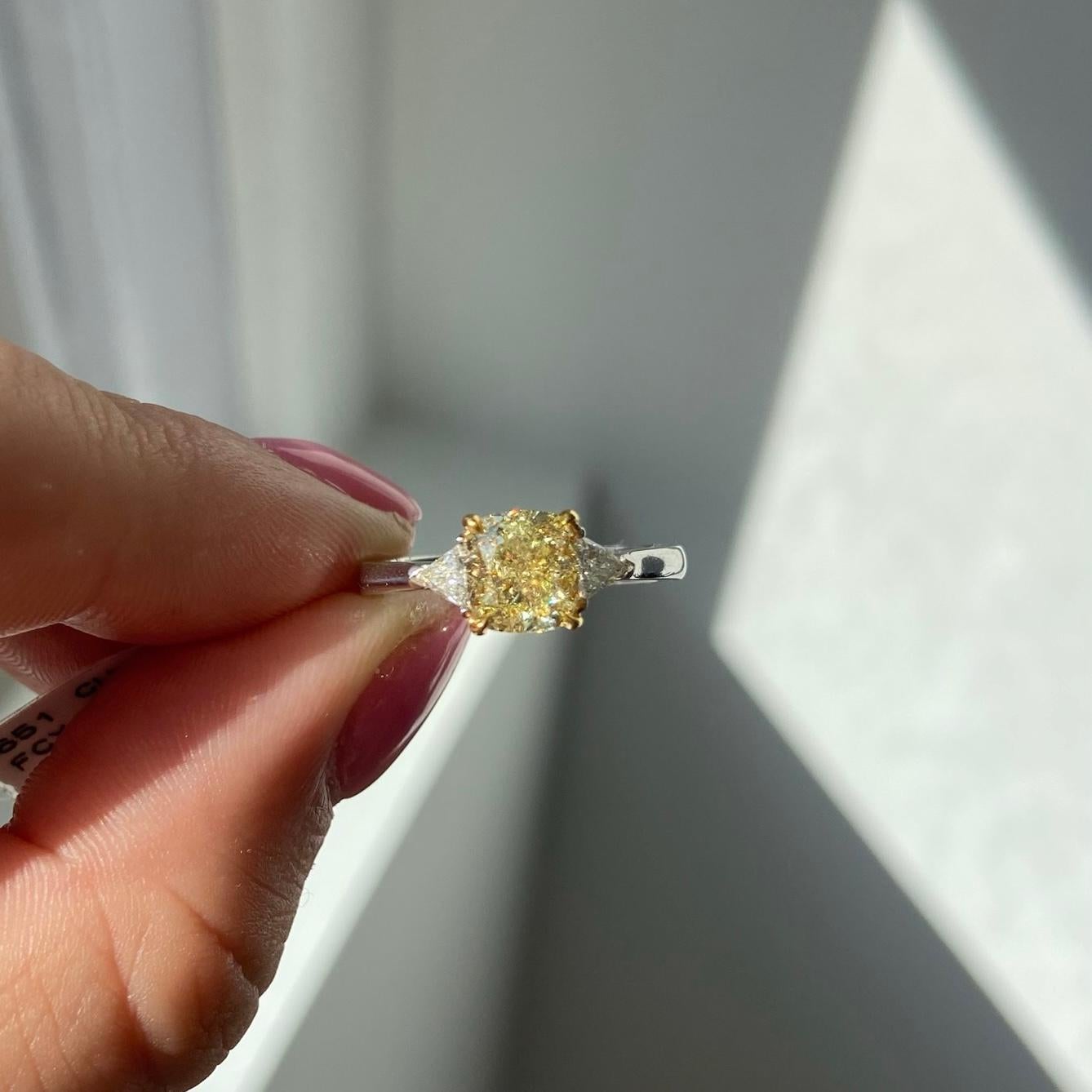 Ein lebendiger Fancy Yellow Cushion Cut Verlobungsring mit drei Steinen, eingefasst in Platin und 18 Karat Gelbgold im Korb, um die natürliche kanariengelbe Farbe voll zur Geltung zu bringen. 

Dieser Diamant hat eine kristallklare Reinheit von VS1