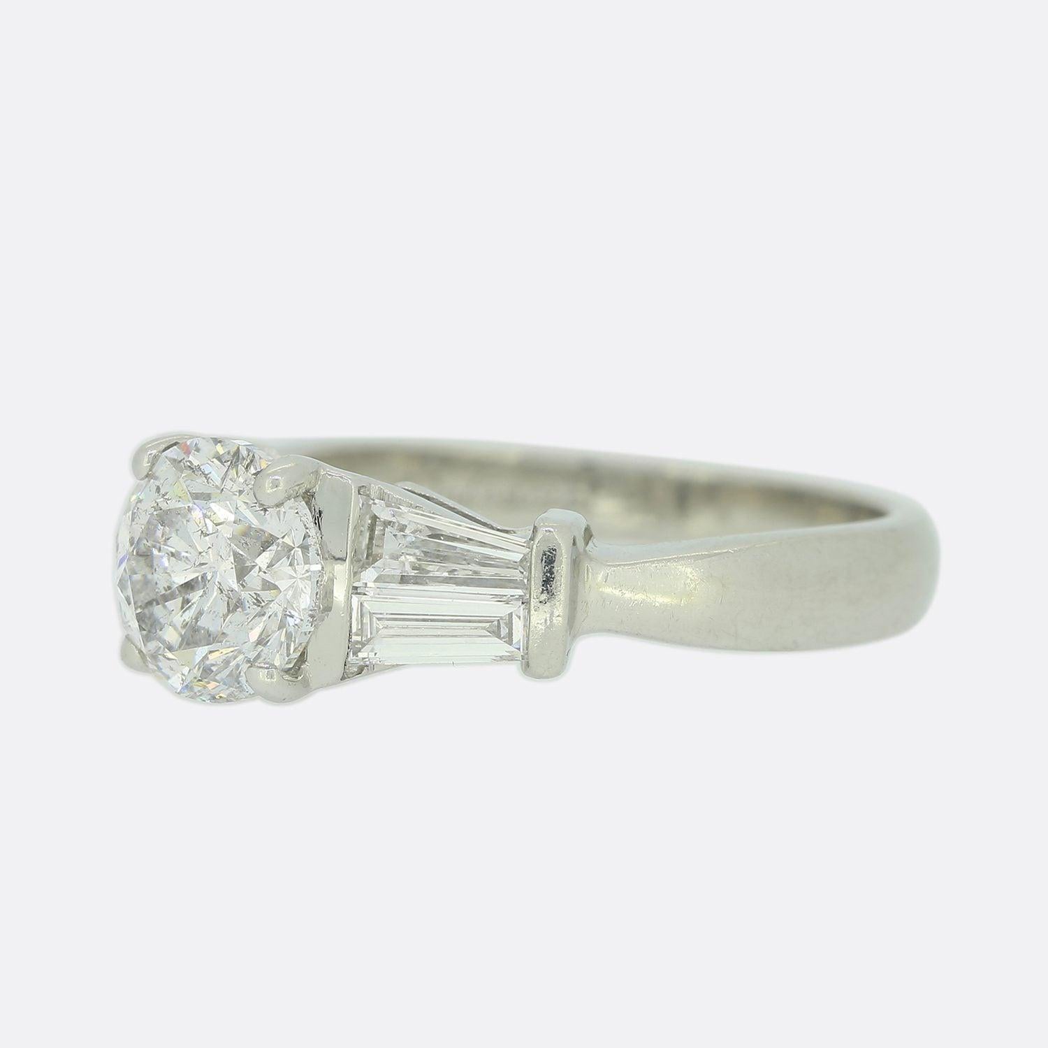 Il s'agit d'une bague de fiançailles en diamant platine. La bague est ornée d'un diamant rond de taille brillant surélevé et de deux diamants baguettes effilés sur chaque épaule. Il porte la marque de Dev Valencia, un créateur de bijoux fins