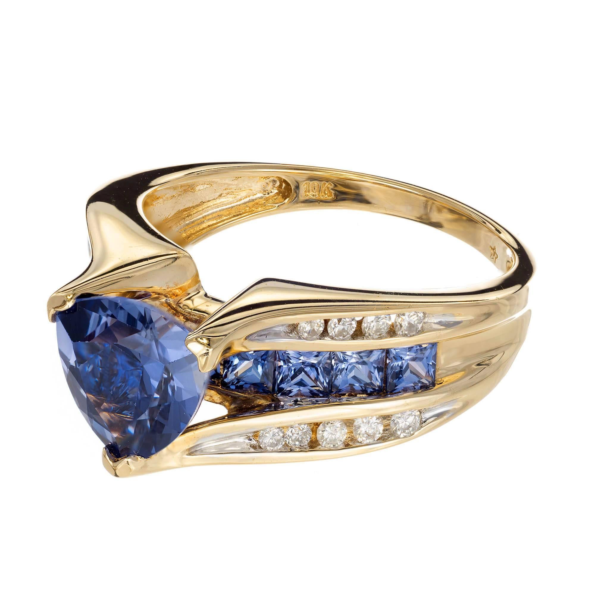 Verlobungsring aus Gelbgold mit Tansanit und Diamant. Abgerundete Trillant-Tanzanite, hoch über einer Reihe von Tansaniten im Prinzessinnenschliff. Eine Reihe runder Diamanten ist in Kanäle gefasst, die zu jeder Zacke führen, die den Haupttansanit