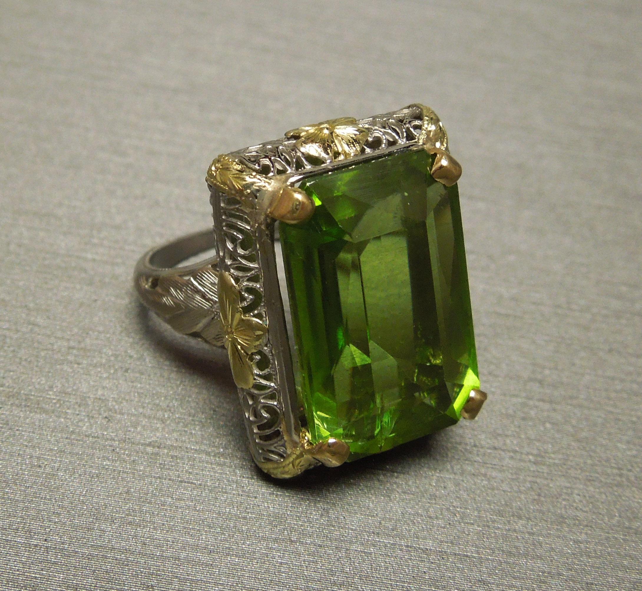 This Antique Peridot Solitaire ring features a GIA Certified Emerald cut 15.40 carat Peridot at 18mm x 11.5mm is securely set in four 18KT Yellow Gold prongs in an intricate Filigree 18KT White, Green & Yellow Gold mounting. [The most desired