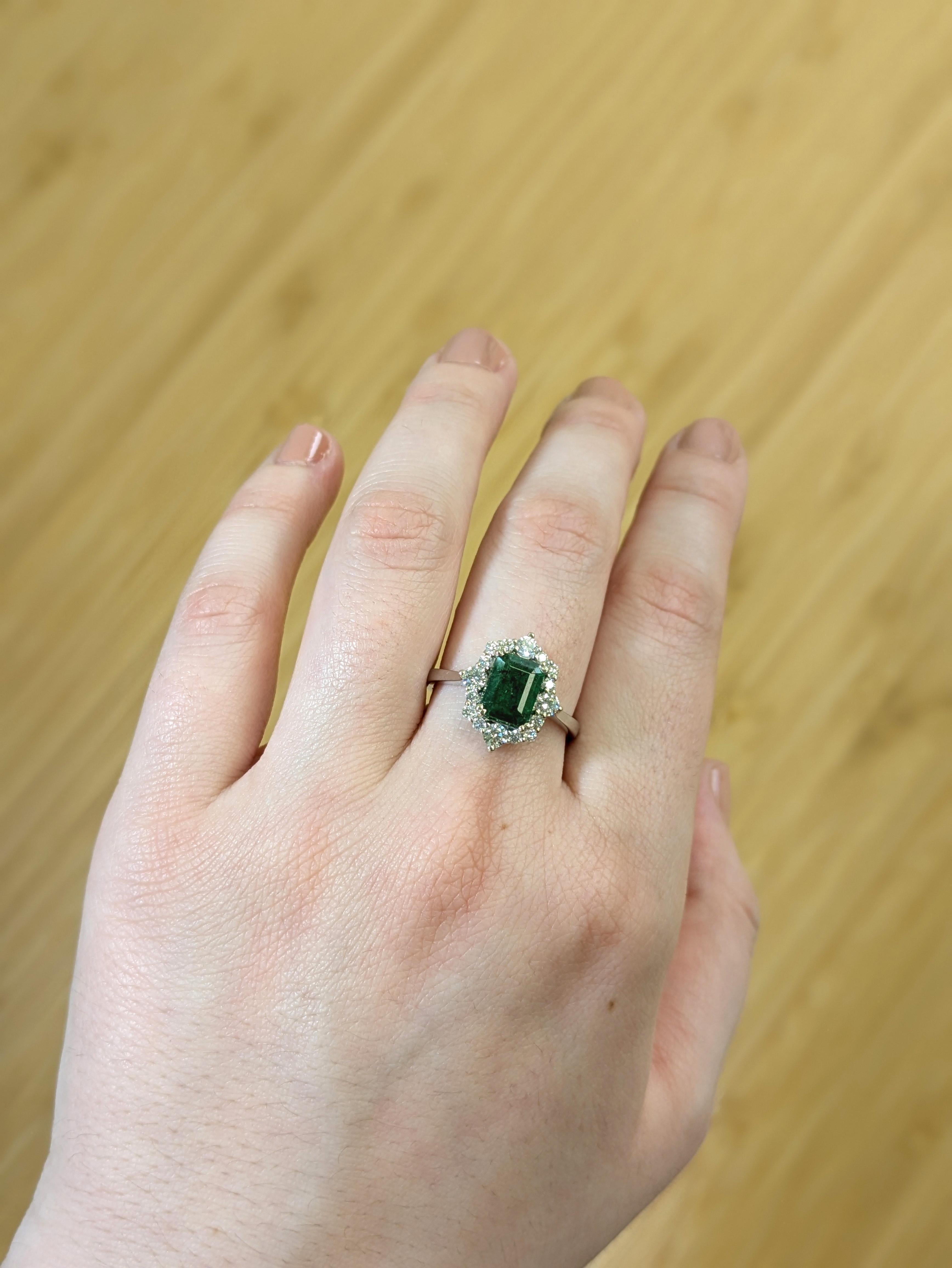 This refined statement ring features a vivid 1.5 carat Zambian emerald, emerald-cut to showcase its natural depth and striking green hue. A halo of sparkling, earth-mined diamonds gently surrounds the center stone, all set in cool, polished 14k