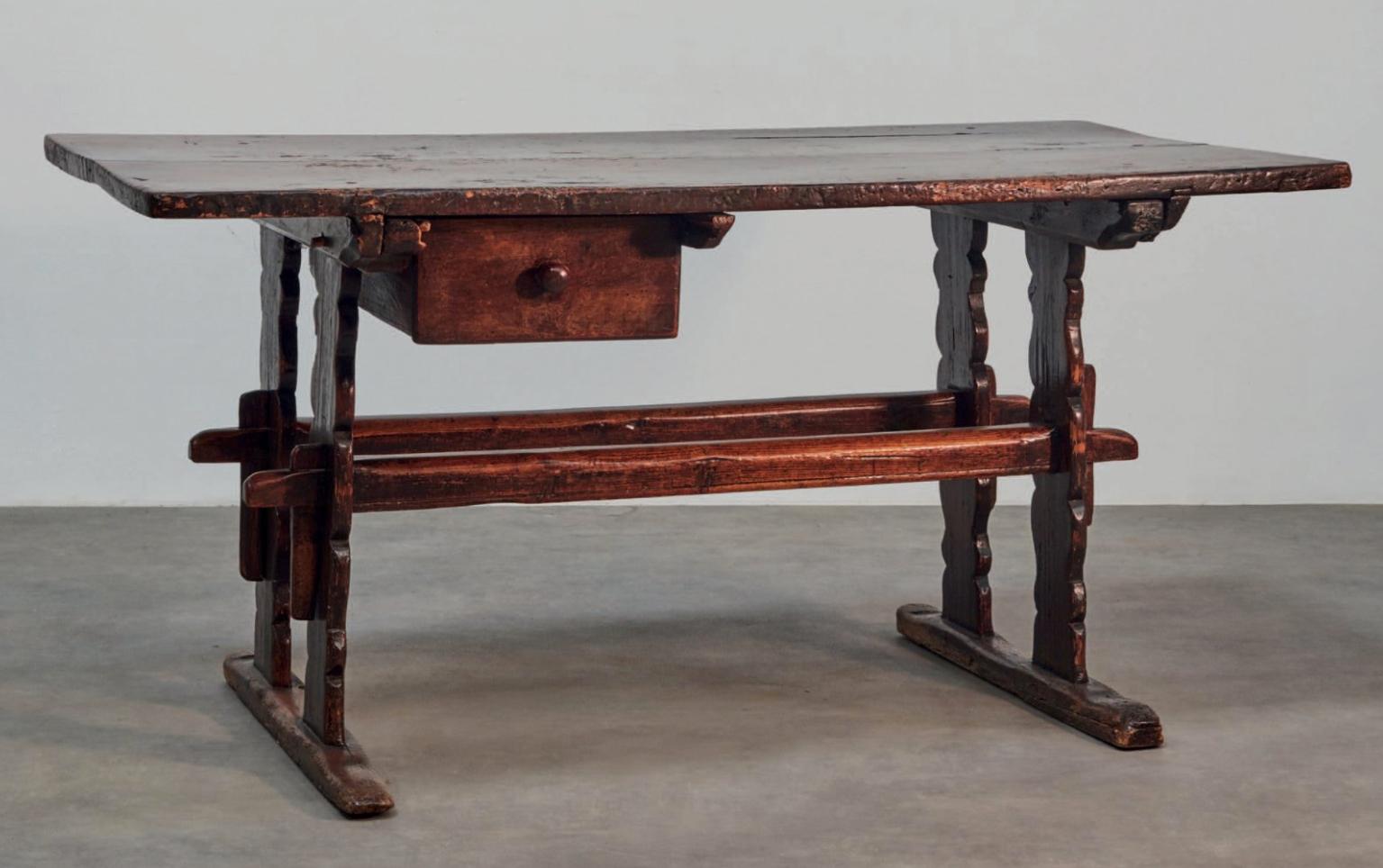 Questo tavolo in noce e legno dolce si regge su due guide. I suoi piedi tagliati sono collegati da distanziatori paralleli montati. L'ampio piano sovrapposto nasconde un cassetto sospeso con pomello in legno tornito.