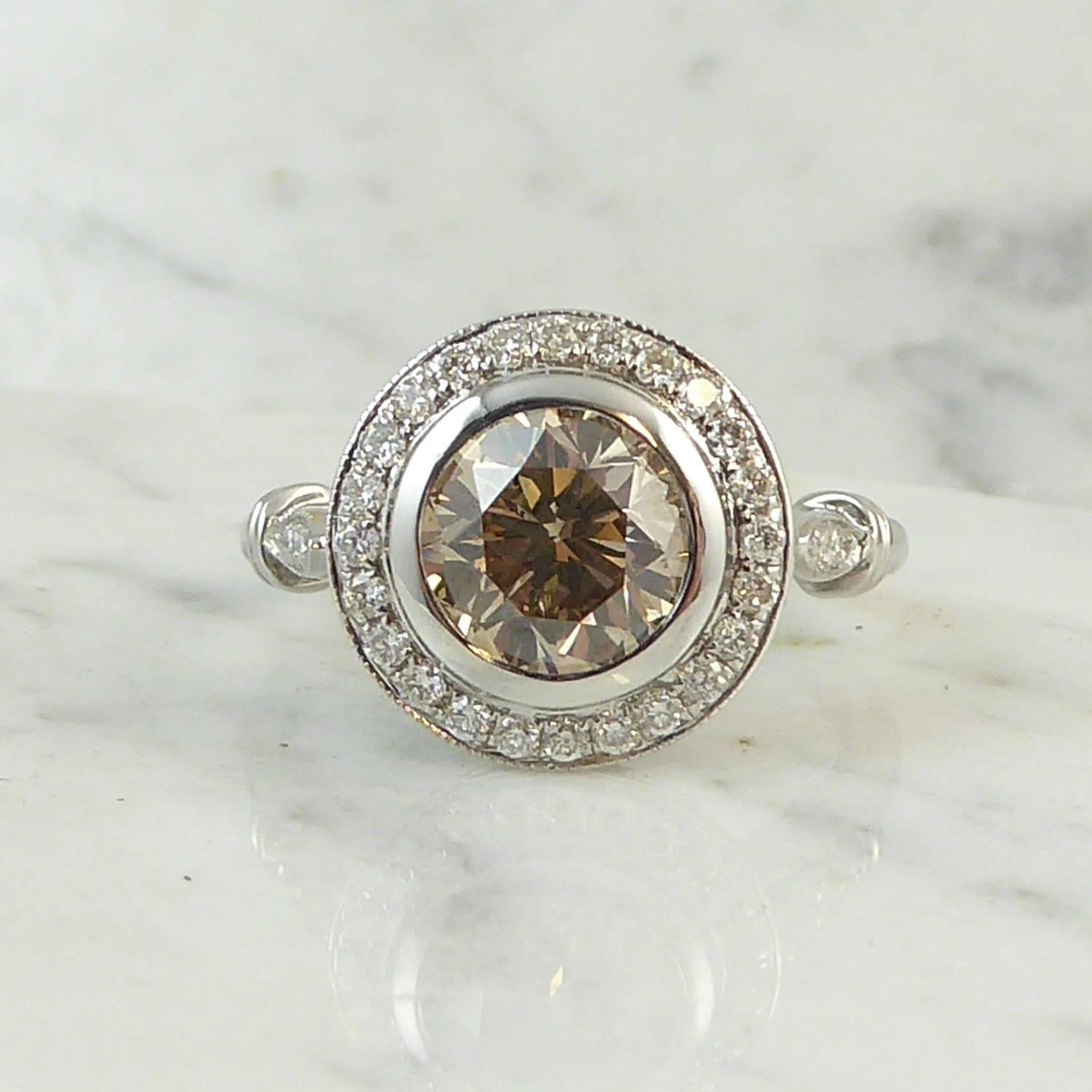 A fancy light yellowish brown diamond set to the centre of a halo of white  diamonds creates a very pleasing colour contrast to this cluster ring.  The fancy diamond is in a white polished rub-over setting to the halo surround of 28 grain set white