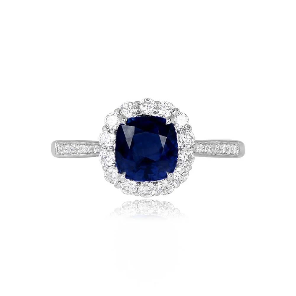 This ring showcases a prong-set cushion-cut sapphire weighing 1.62 carats. Surrounding the center sapphire is a halo of round brilliant-cut diamonds, with additional micro-pave-set diamonds on the shoulders. The total diamond weight of this
