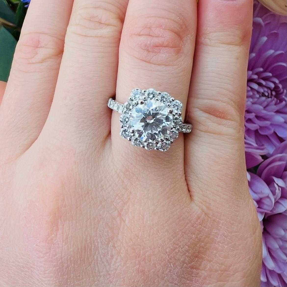 A gorgeous ring design featuring a fantastic center stone! This 18 karat white gold beauty features at its center a 1.62 carat natural round brilliant cut diamond, surrounded by a halo of fine white melee diamonds and additional diamonds set in the