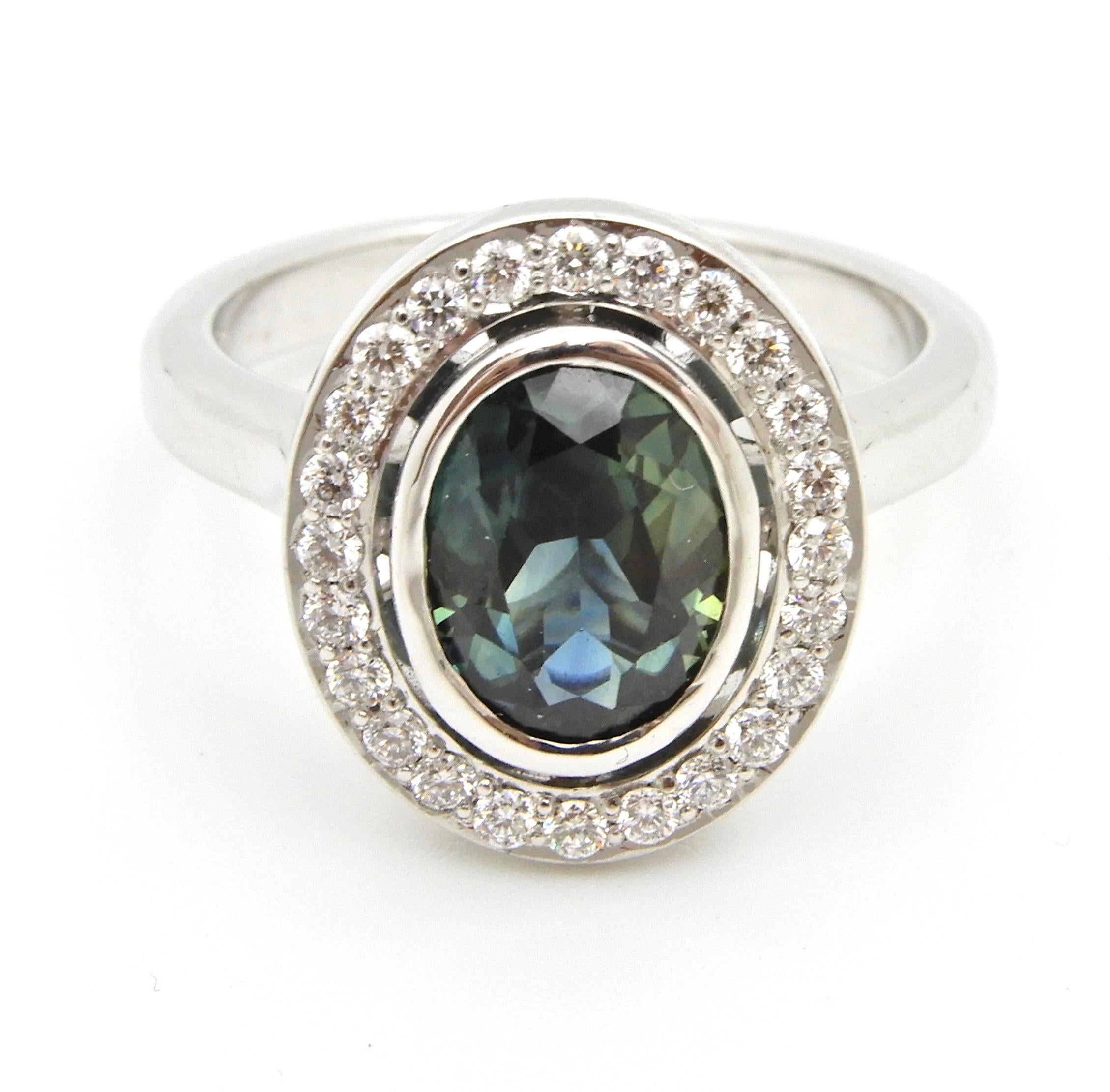 This 1.65 Carat Oval Cut Teal Blue Sapphire and Diamond Engagement Ring has a slightly rounded, flat edge band with rising, pointed shoulders that supports a central, scalloped oval gallery holding the central rub over oval sapphire. The ring