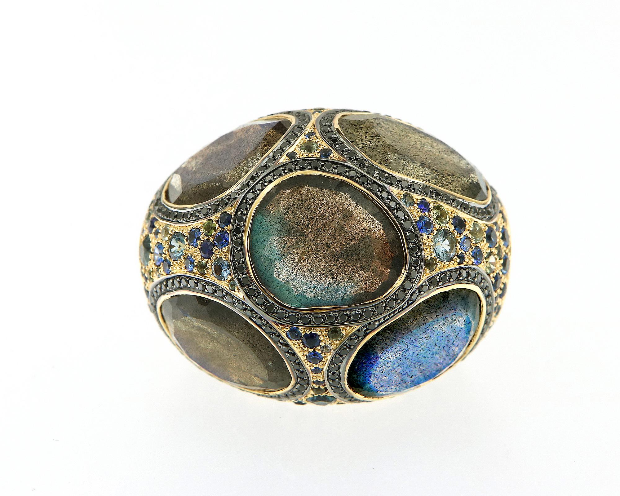 This bohemian glam ring is created with 18k yellow gold and showcases beautiful free form labradorite weighing 16.54 carats in the design. Blue and green sapphires liberally splashed around the surface of the dome, creating a beautiful and sparkling