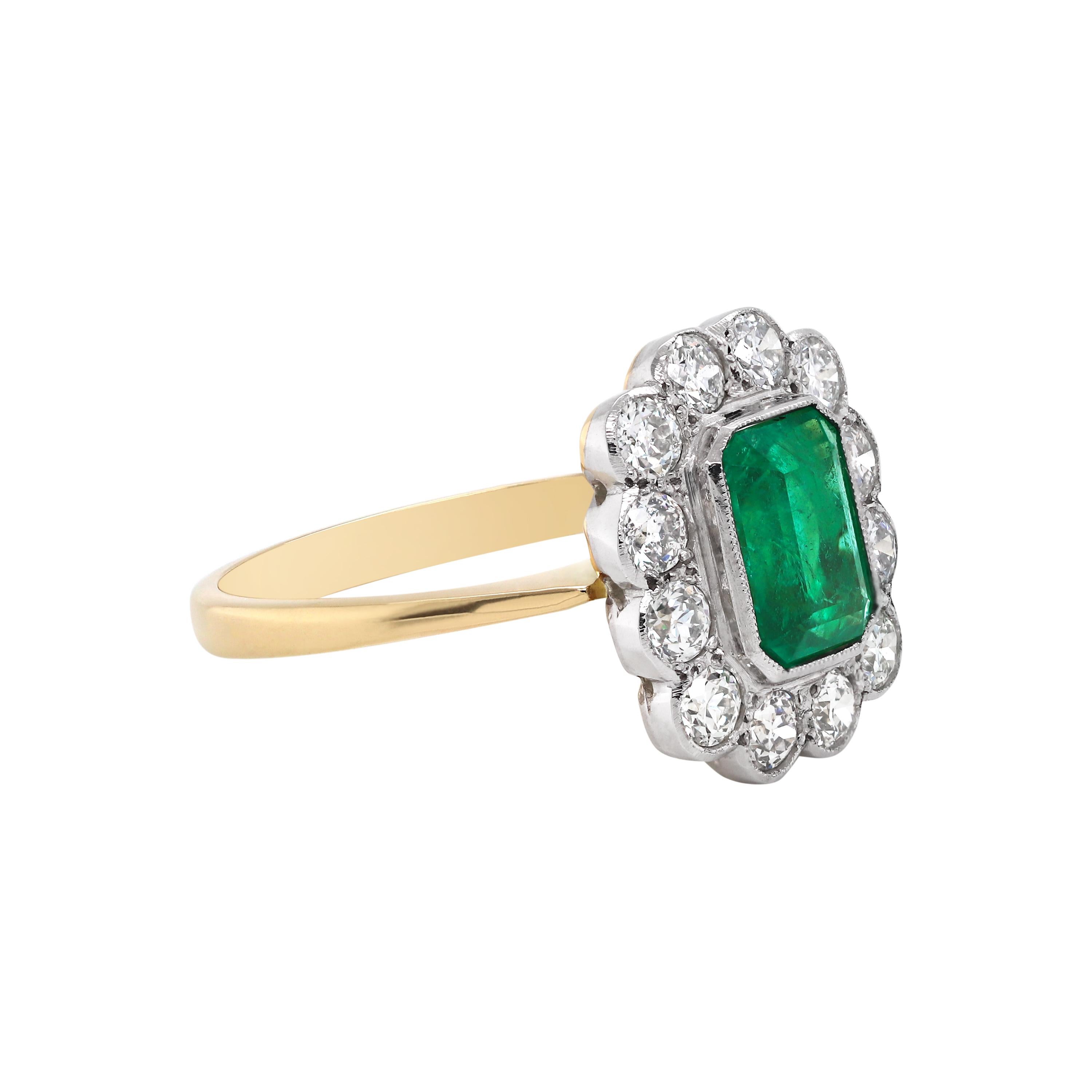 This vintage inspired coronet cluster engagement ring features a vibrant 1.65 carat emerald cut emerald mounted in a rubover, open back setting. The emerald is beautifully surrounded by twelve fine quality round brilliant cut diamonds weighing a