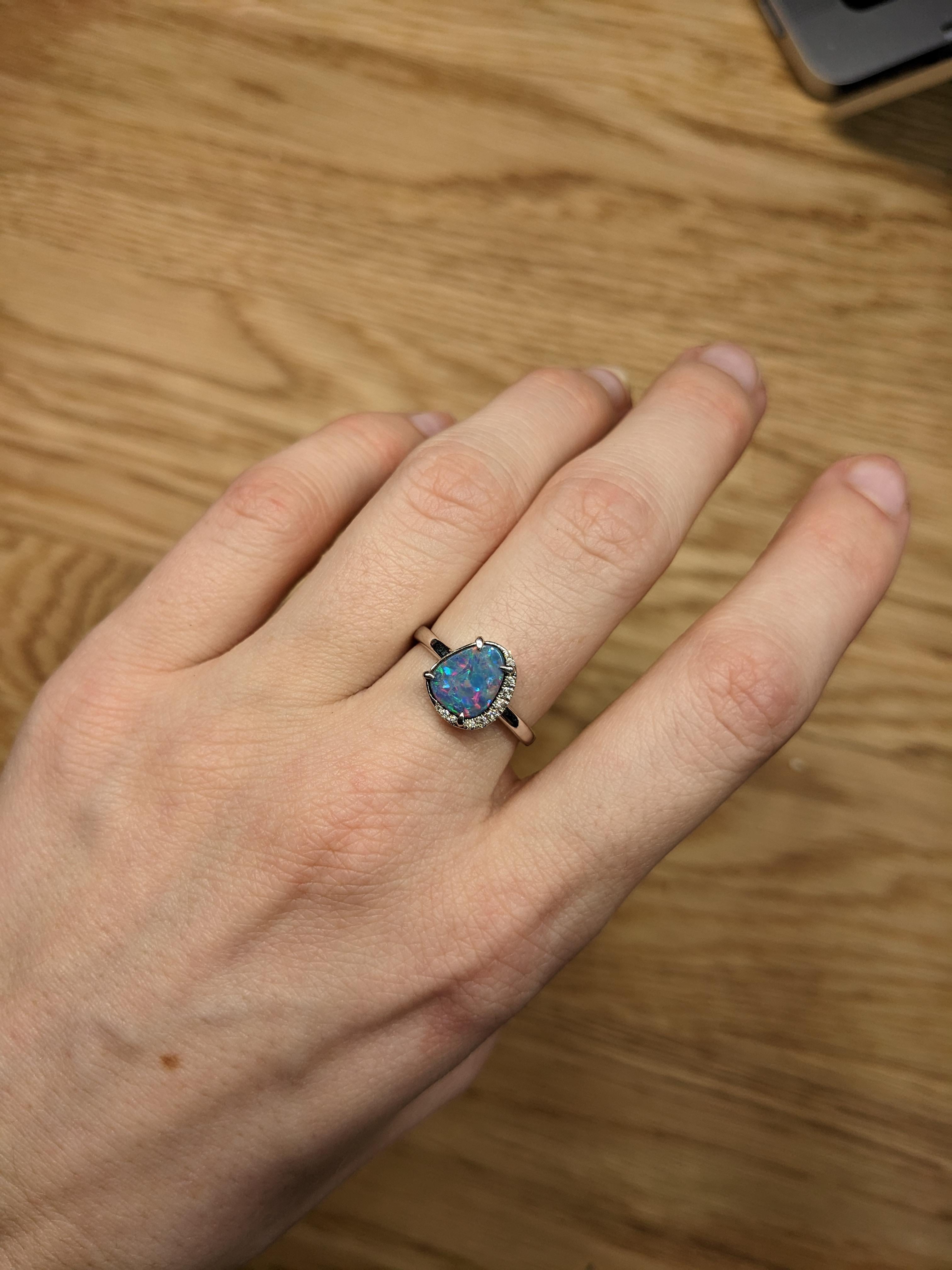 The 14K White Gold 1.65cts Boulder Opal Diamond Ring features a stunning fancy shape 9x7mm Boulder Opal at its center, surrounded by a delicate halo of diamonds. The opal's vibrant, iridescent colors are set in a luxurious 14K white gold band. The