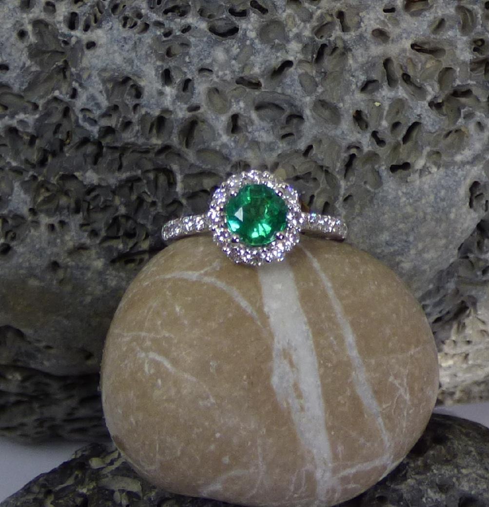 Ein leuchtend bunter Smaragd ist in diesem Cluster-Ring gefasst. Der Smaragd ist 7 mm rund und 1,16ct schwer.  Er ist von 12 Diamanten und 6 Diamanten auf jeder Seite umgeben. Das Gesamtgewicht der Diamanten beträgt 0,70ct.  Der Ring ist