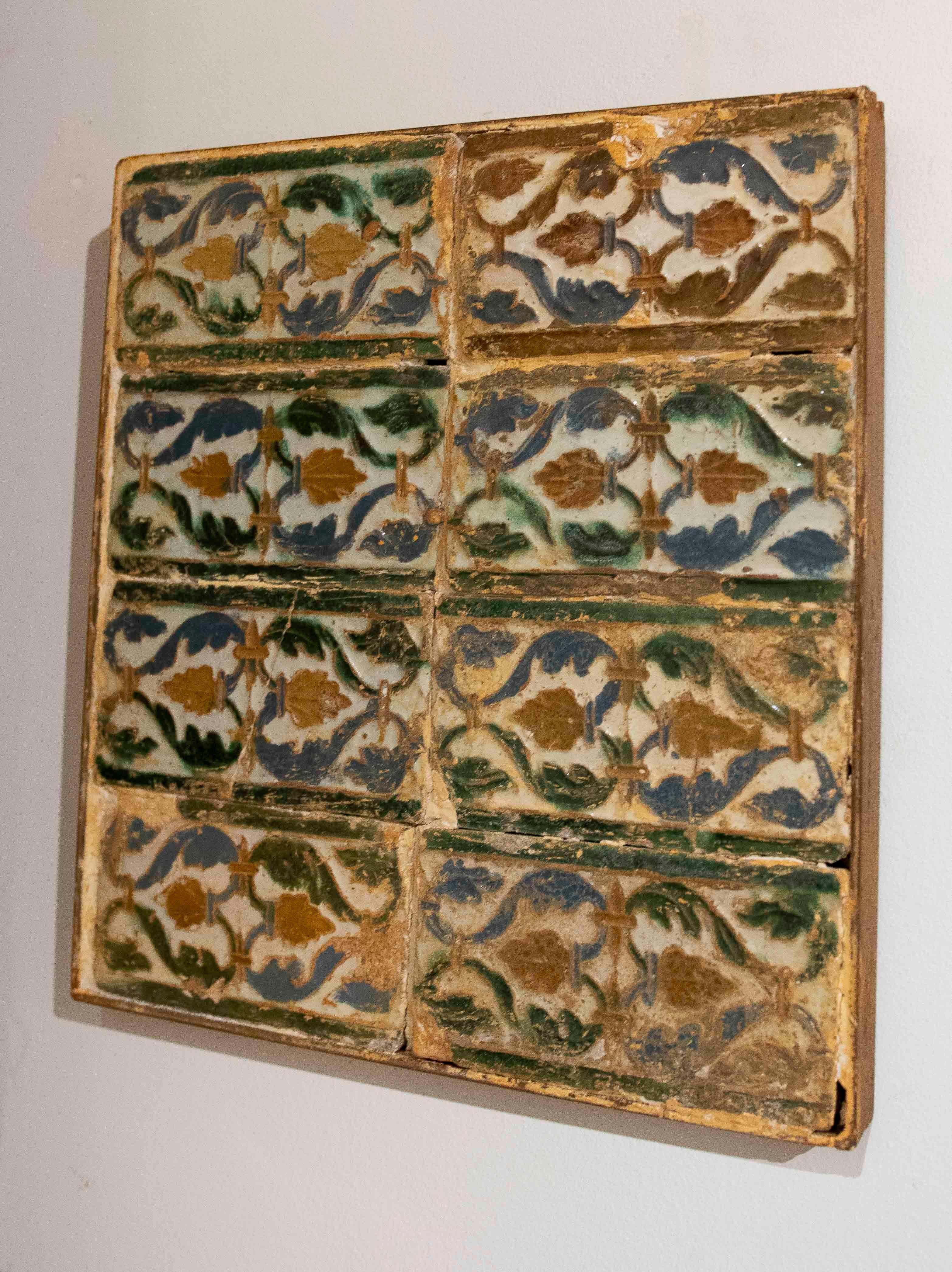 Panel composed of antique rectangular tiles decorated with plant motifs in shades of green, blue, and ochre. Each piece displays patina, craquelure, and natural wear, enhancing its historical and artisanal character. The arrangement offers a