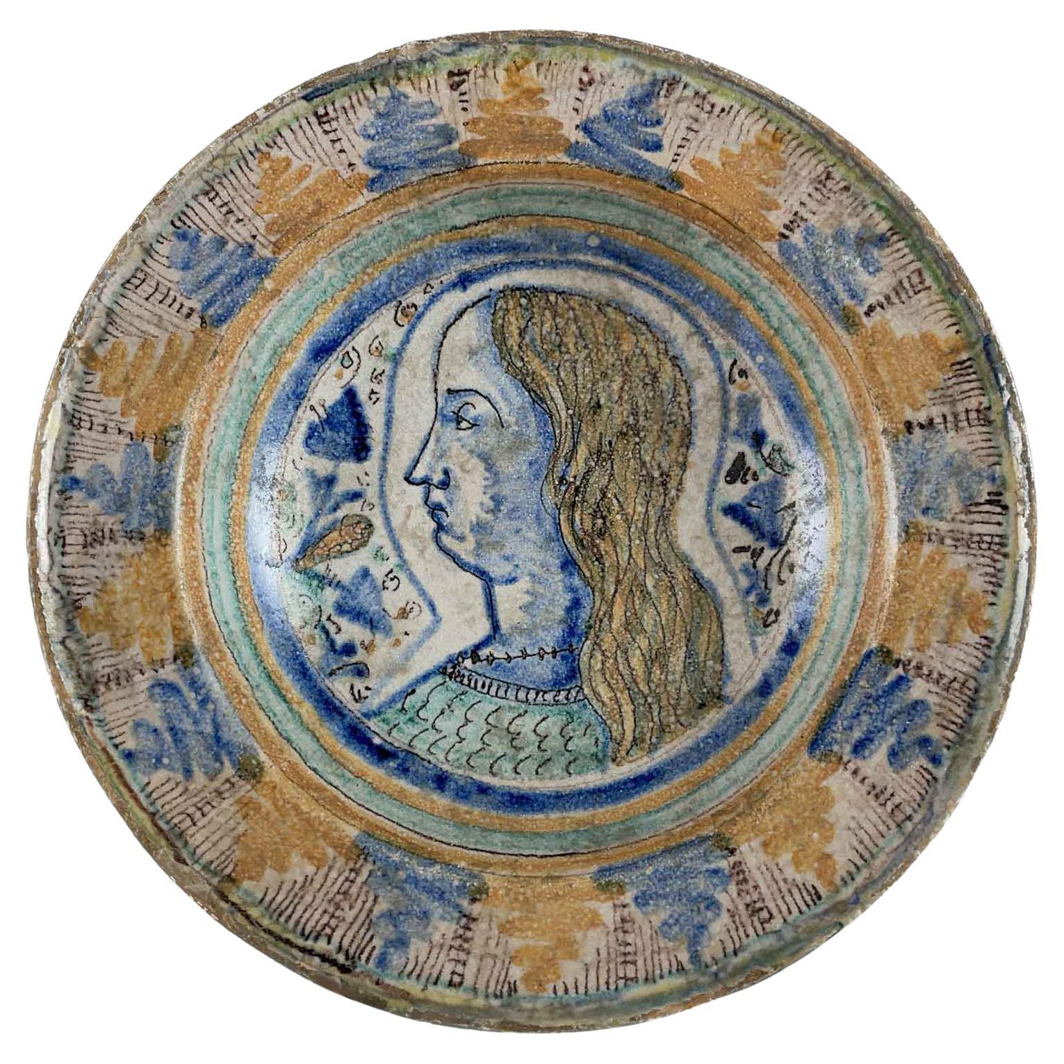 Plat italien en maïolique du milieu du XVIe siècle, peint à la main en bleu et jaune ocre, avec un portrait de jeune homme de profil au centre. Les couleurs jaunâtres et bleues de la maïolique sont peintes dans une élégante alternance de motifs